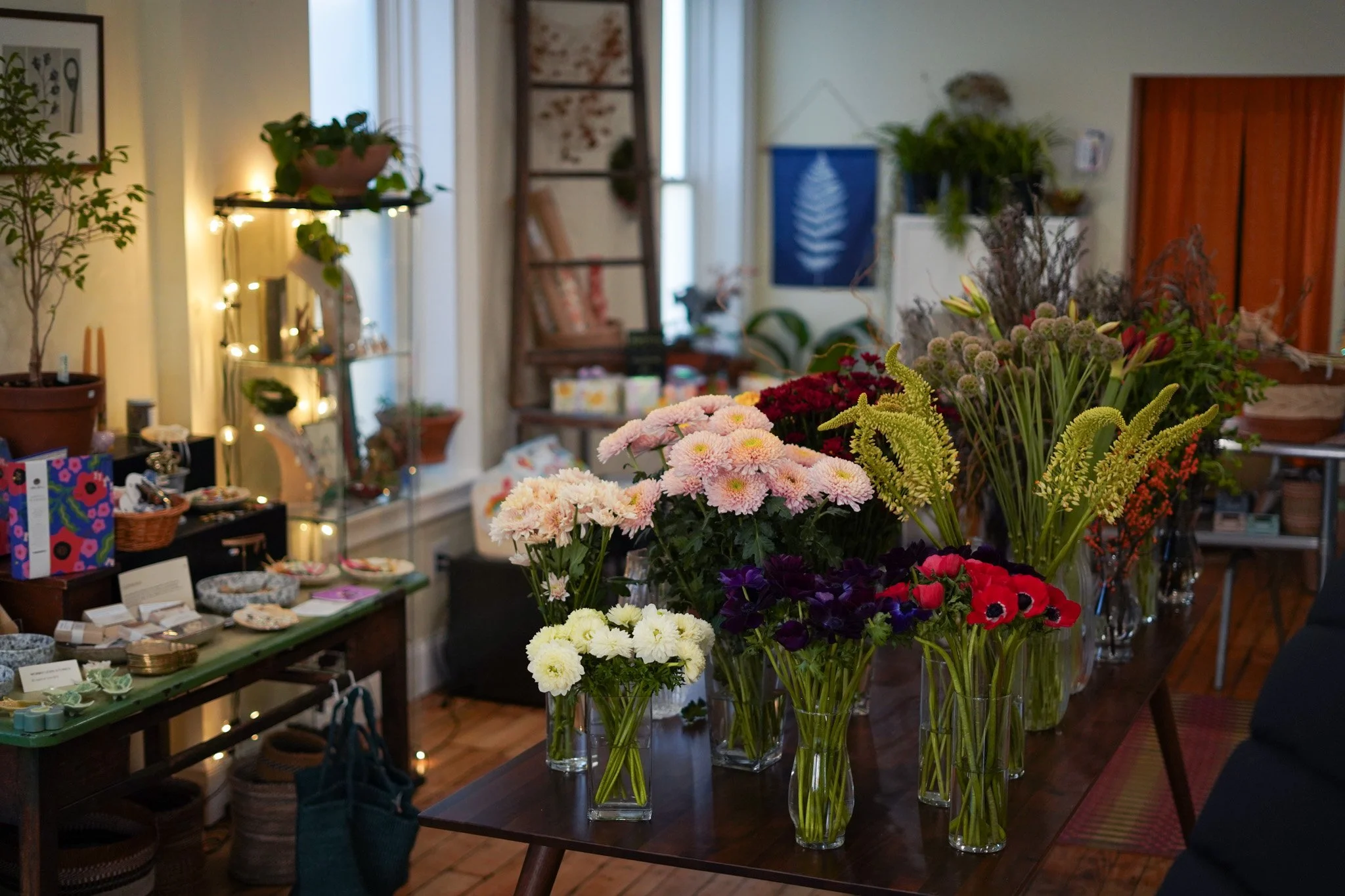 A large wooden table with many colorful flowers in glass vases, and shelves displaying various items and potted plants in a cozy, well-decorated room.
