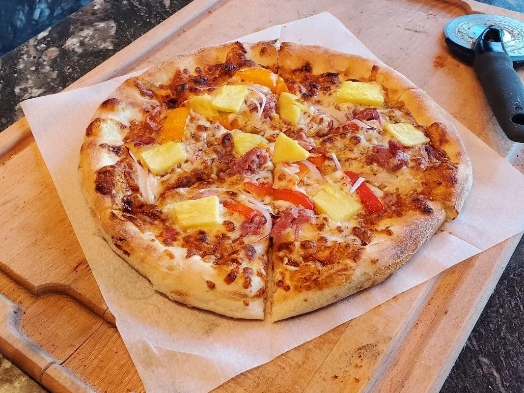 Pizza with pineapple, bell pepper, ham, and cheese on a wooden board with a pizza cutter.