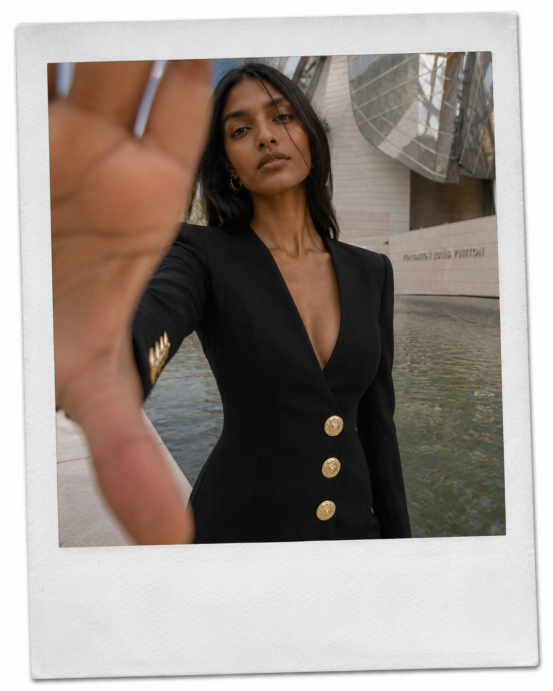 A woman taking a selfie outdoors by a modern building with water, wearing a black blazer with gold buttons, with her hand extended toward the camera.