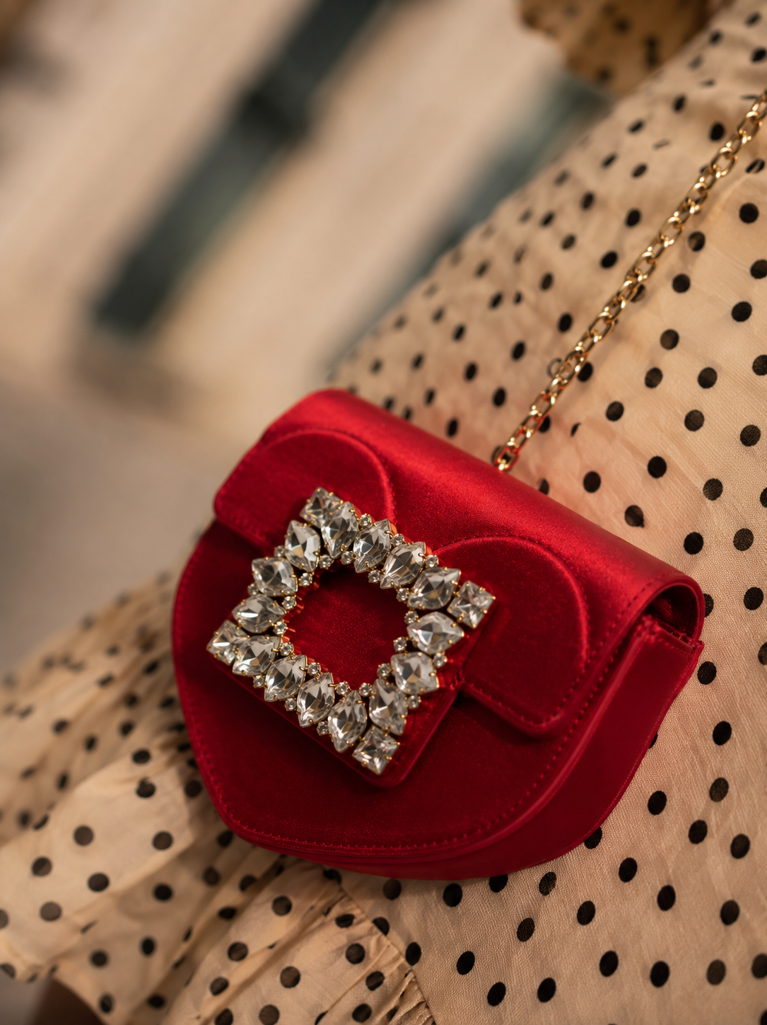A red satin clutch purse with a large square rhinestone embellishment on the front, resting on a polka dot fabric with a beige background and black dots, accompanied by a gold chain strap.
