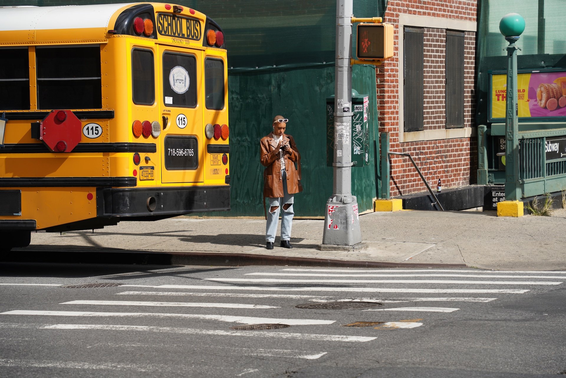 A yellow school bus parked on the street with a woman standing nearby, looking at her phone. She is wearing a brown leather jacket, ripped jeans, and sunglasses. There are street signs, a brick building, and a Subway sign in the background.