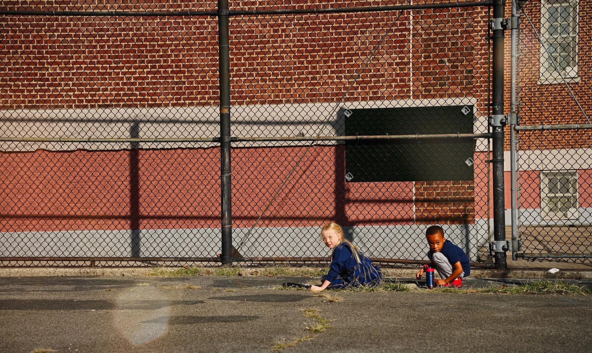 Two children sitting on the sidewalk in front of a chain-link fence with a red brick building in the background.
