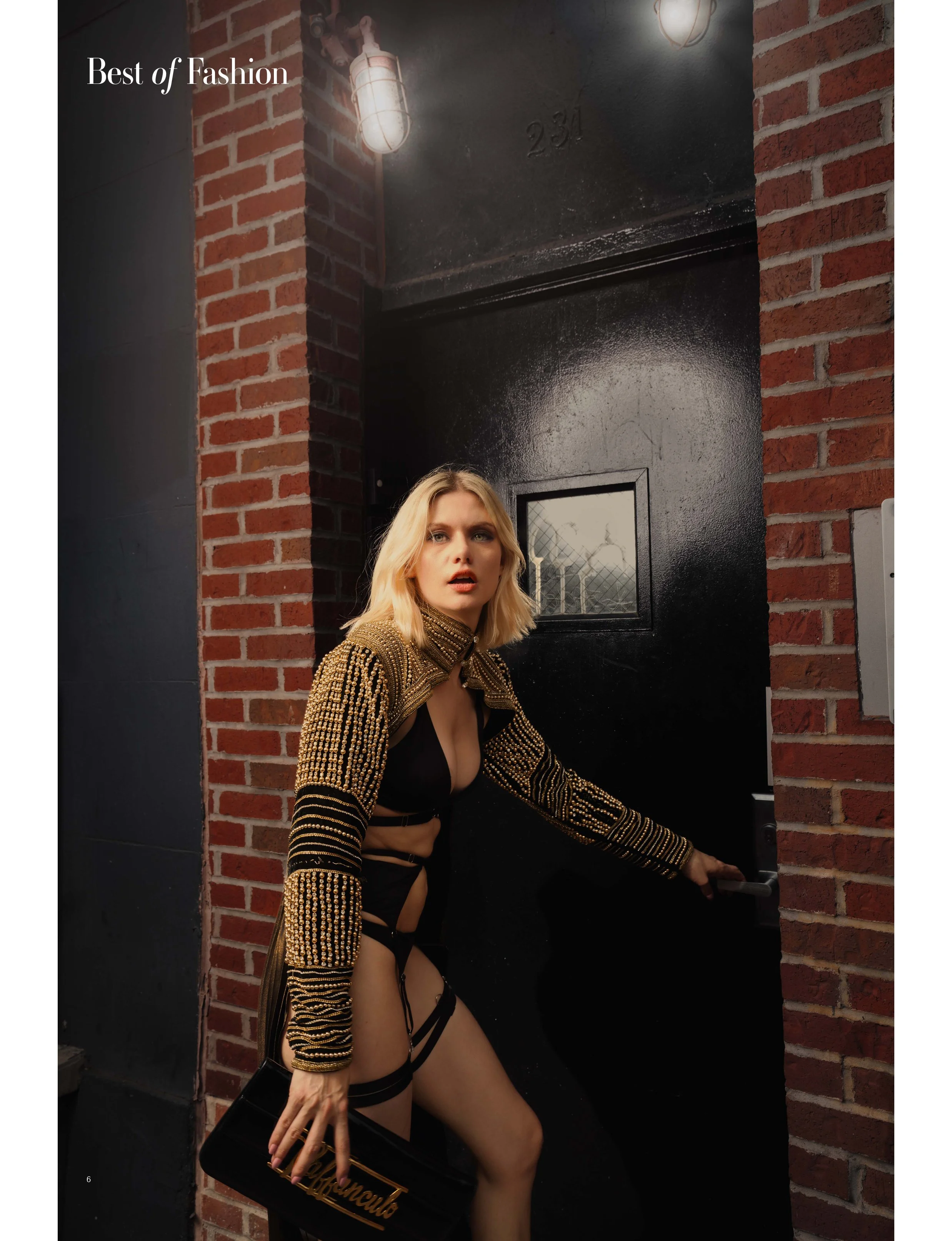Fashion model wearing black and gold embellished outfit holding a purse, standing by a brick wall next to a black door marked 231, illuminated by overhead lights.