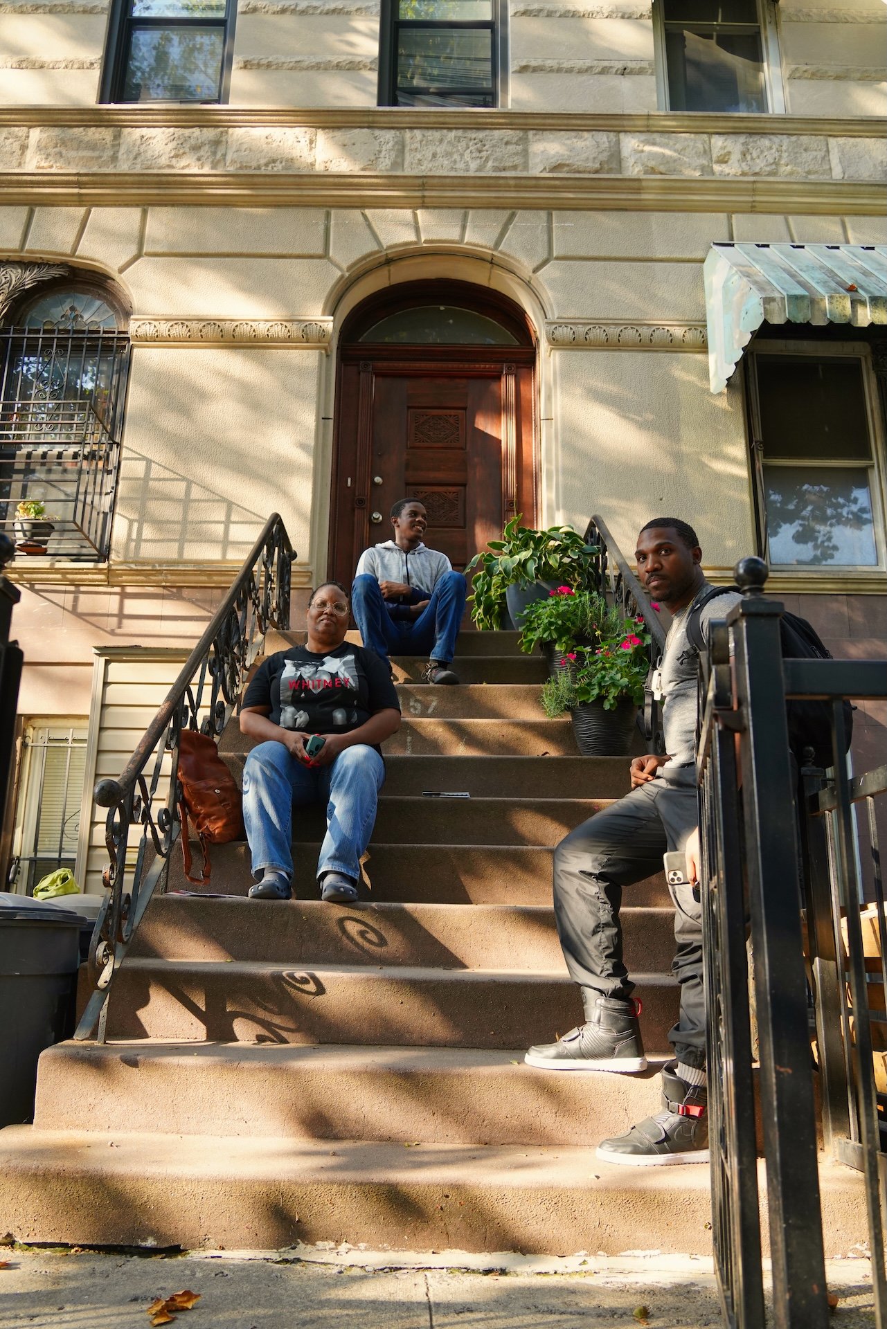 Three people sitting on steps in front of a residential building, with potted plants and shadows cast on the wall.