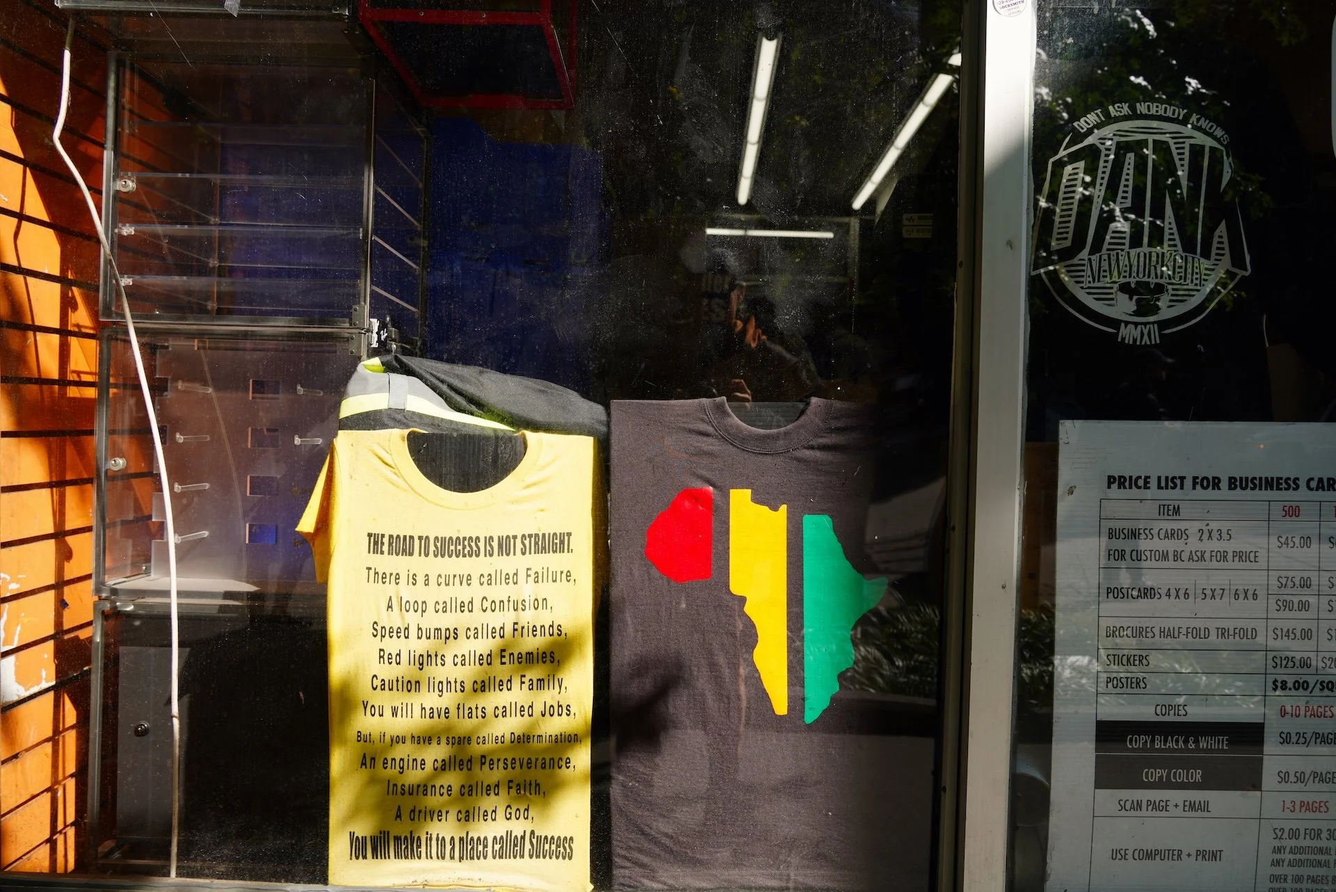 Display case with T-shirts and promotional posters, including a yellow T-shirt with an motivational quote, and a black T-shirt with a colorful map design. There is a logo on the window for a New York City-based brand and a business price list on the right.