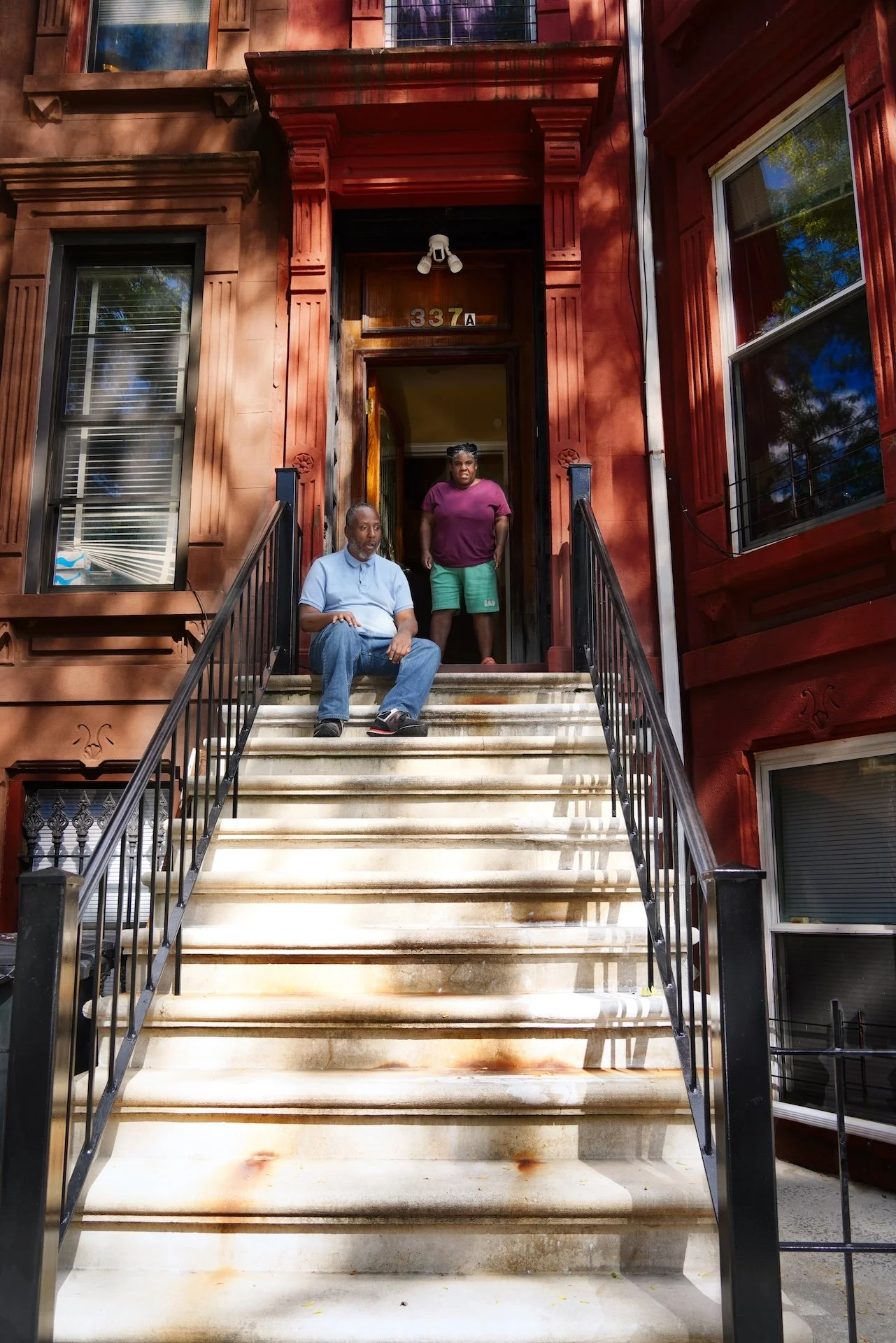 A man sitting on a stairway outside a brownstone building, with a woman standing at the top of the stairs in the doorway. The building has the numbers 337A above the door, and the stairway has black railings on each side.
