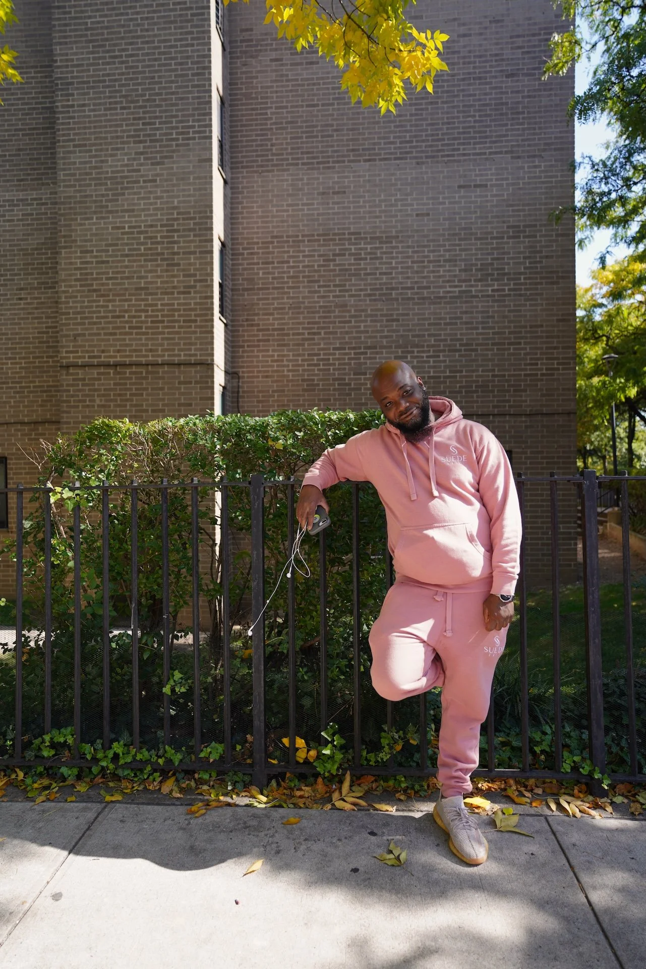 A man in a pink hoodie and sweatpants leaning against a black metal fence, outdoors on a sidewalk with fallen leaves. Behind him are bushes and a brick building, with trees and blue sky visible.