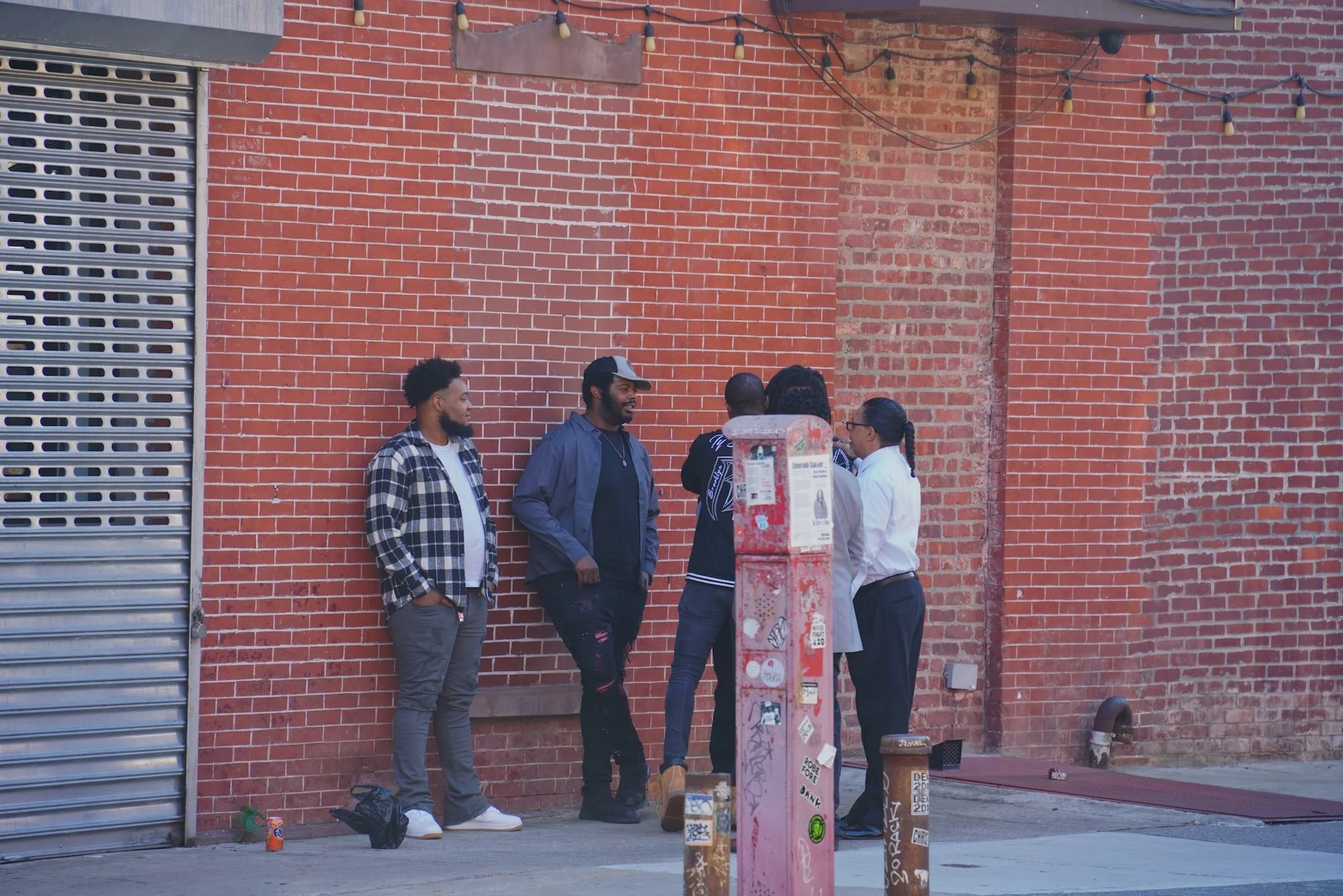 Five people standing and chatting outside against a red brick wall, with a metal pole covered in stickers and graffiti in the foreground.