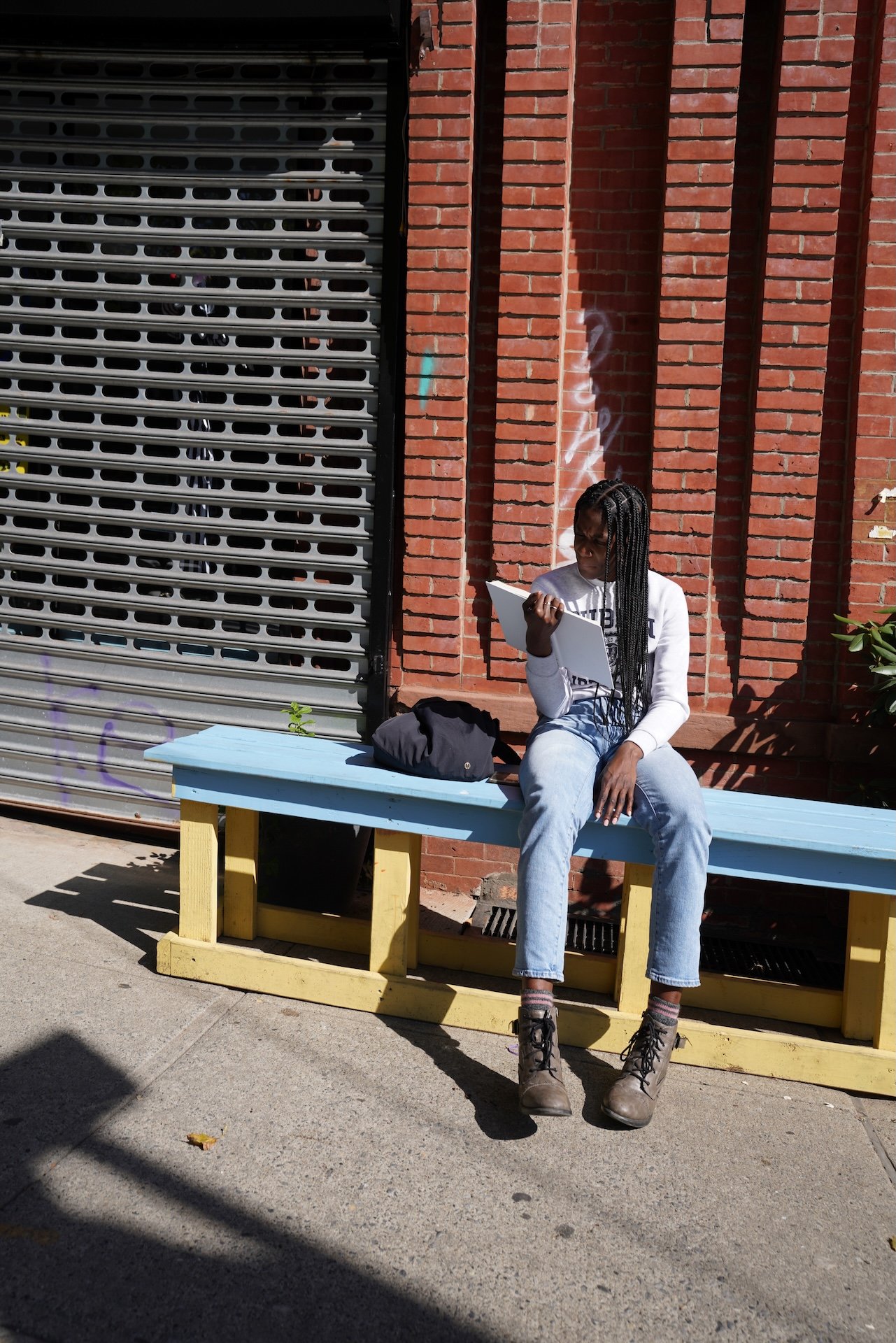 A woman with long braided hair sitting on a light blue and yellow bench outside, reading a book or notebook, with a black bag beside her. She is wearing a white sweatshirt, light blue jeans, and brown boots. The background features a brick wall and a metal rolling shutter.
