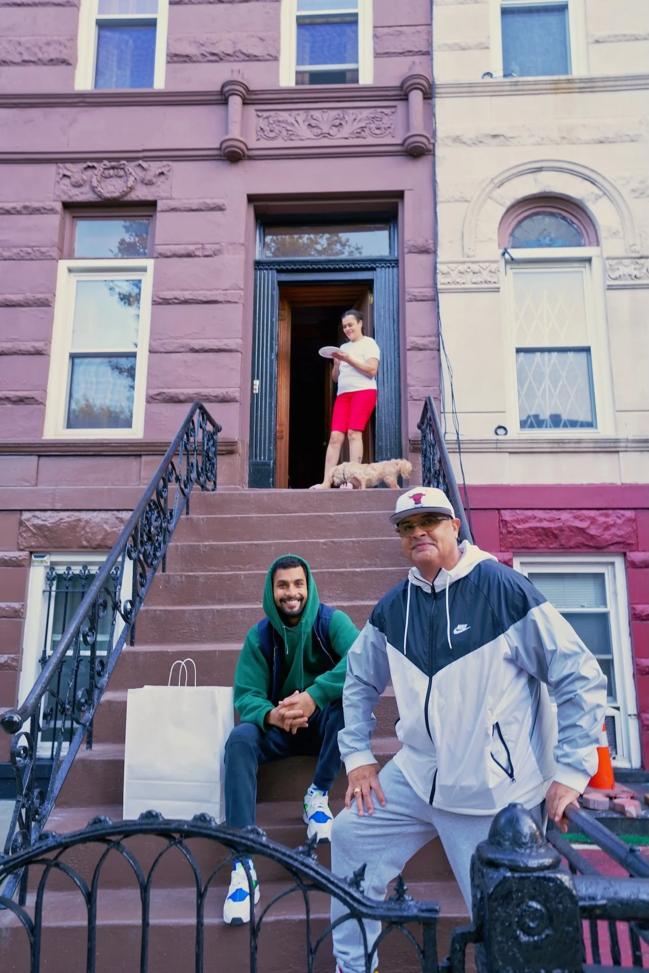 People sitting and standing on the steps of a brownstone house with ornate window frames and decorations, two men in the foreground, a woman at the top of the steps, and a small dog near the woman.