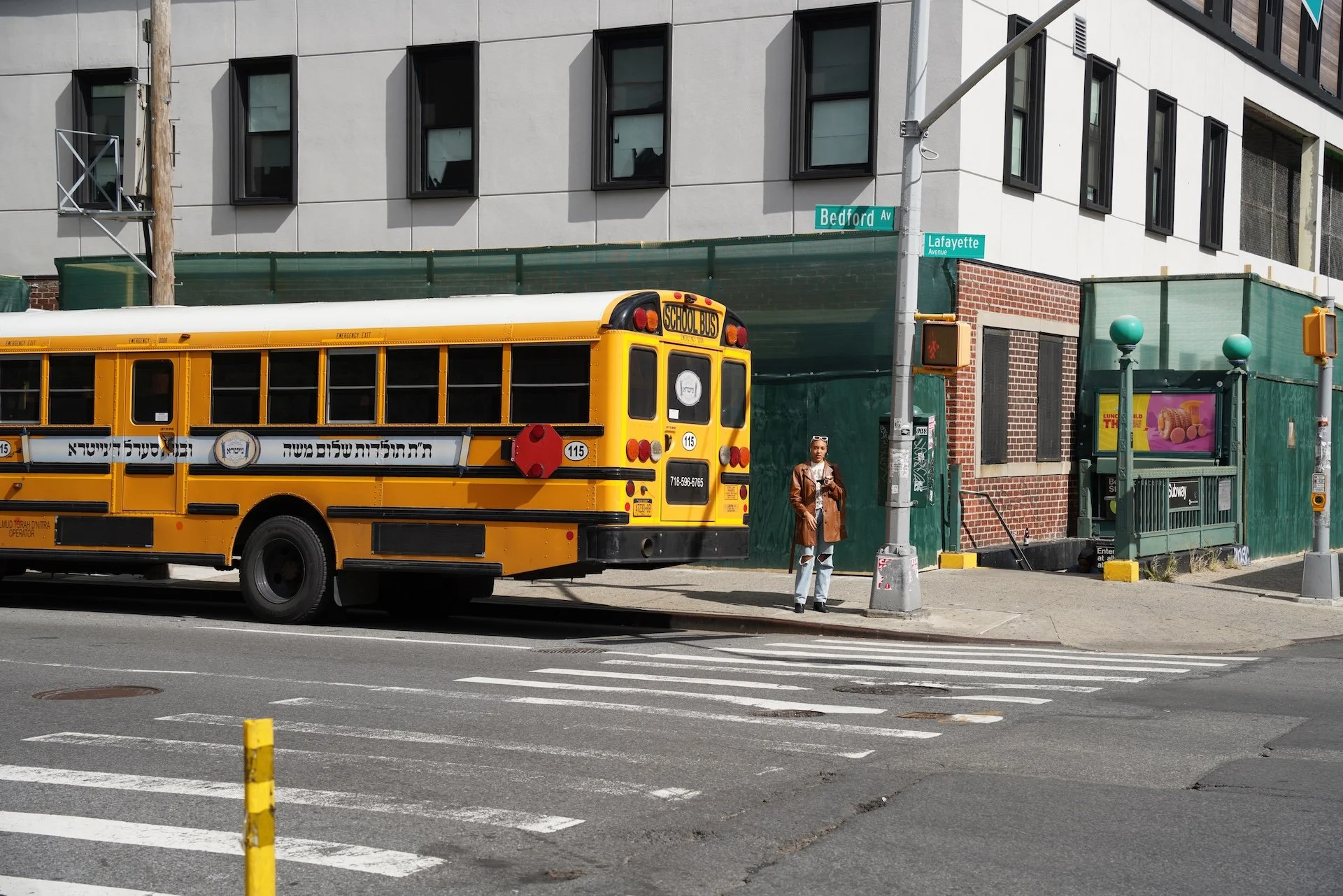 A woman standing at a crosswalk on a city street near a yellow school bus parked by the sidewalk. The background features a modern building with rectangular windows and street signs indicating Bedford Avenue and Lafayette Avenue.