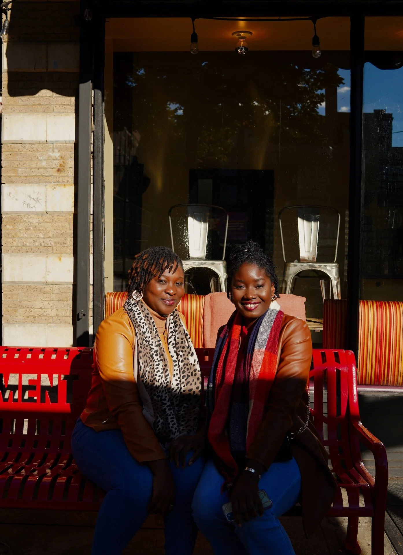 Two women sitting together on a red bench outside a building, smiling. One woman has short, dreadlocked hair and a leopard print scarf, the other has curly hair and a colorful plaid scarf. The background shows a large glass window with chairs and trees reflected in it.