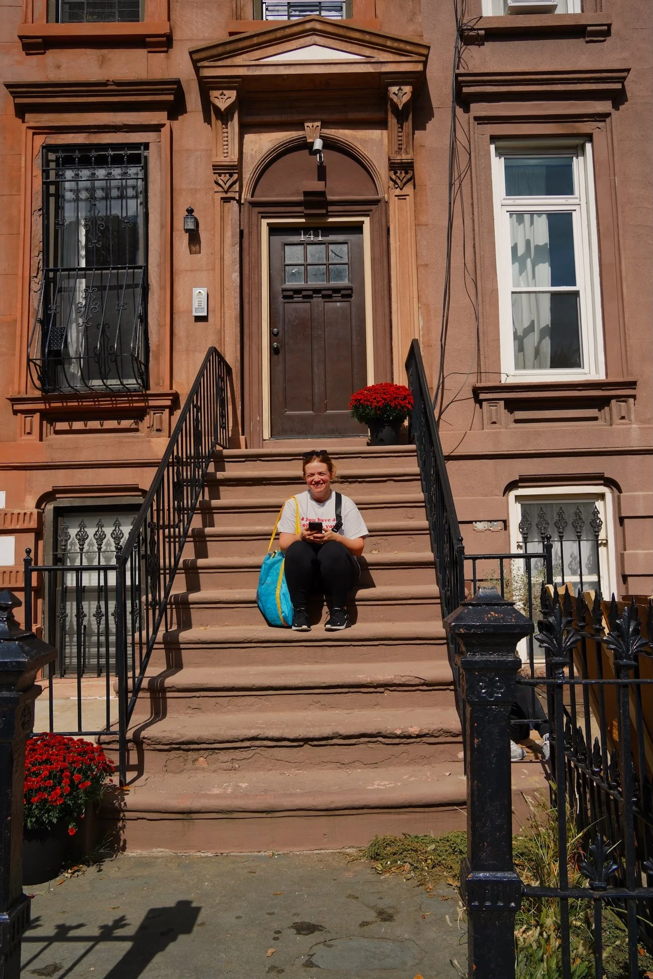 A woman sitting on the steps of a brownstone house, smiling and looking at her phone. She has a yellow and blue backpack next to her. The house has a front door with the number 141, two windows with decorative trim, and a potted red chrysanthemum plant on the porch. Black metal railings line the stairs.