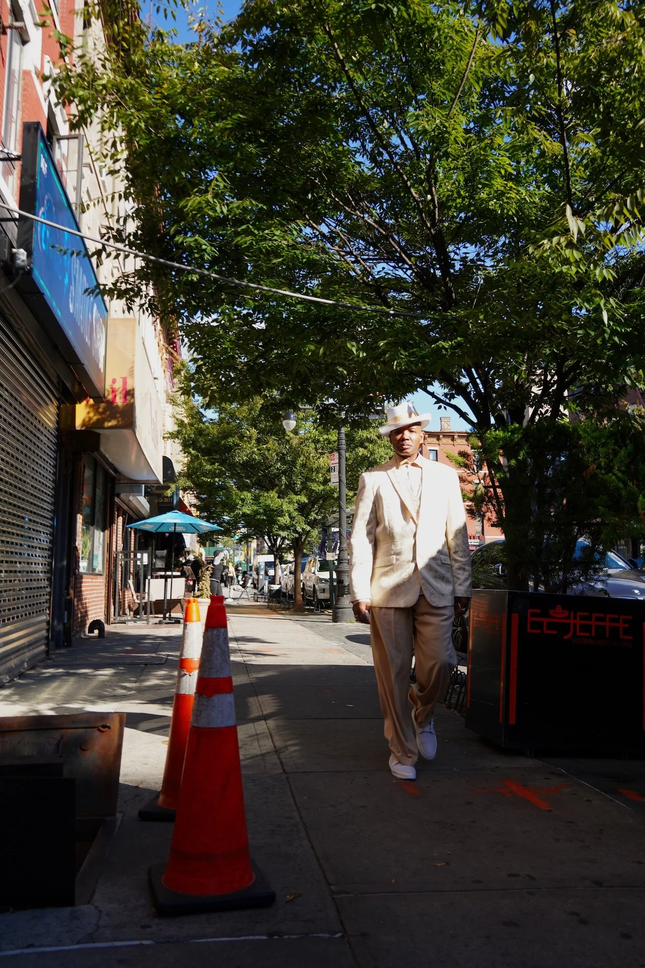 Man in a beige suit walking on a city sidewalk under a tree with green leaves, wearing a white hat, in an urban area with shops and parked cars.