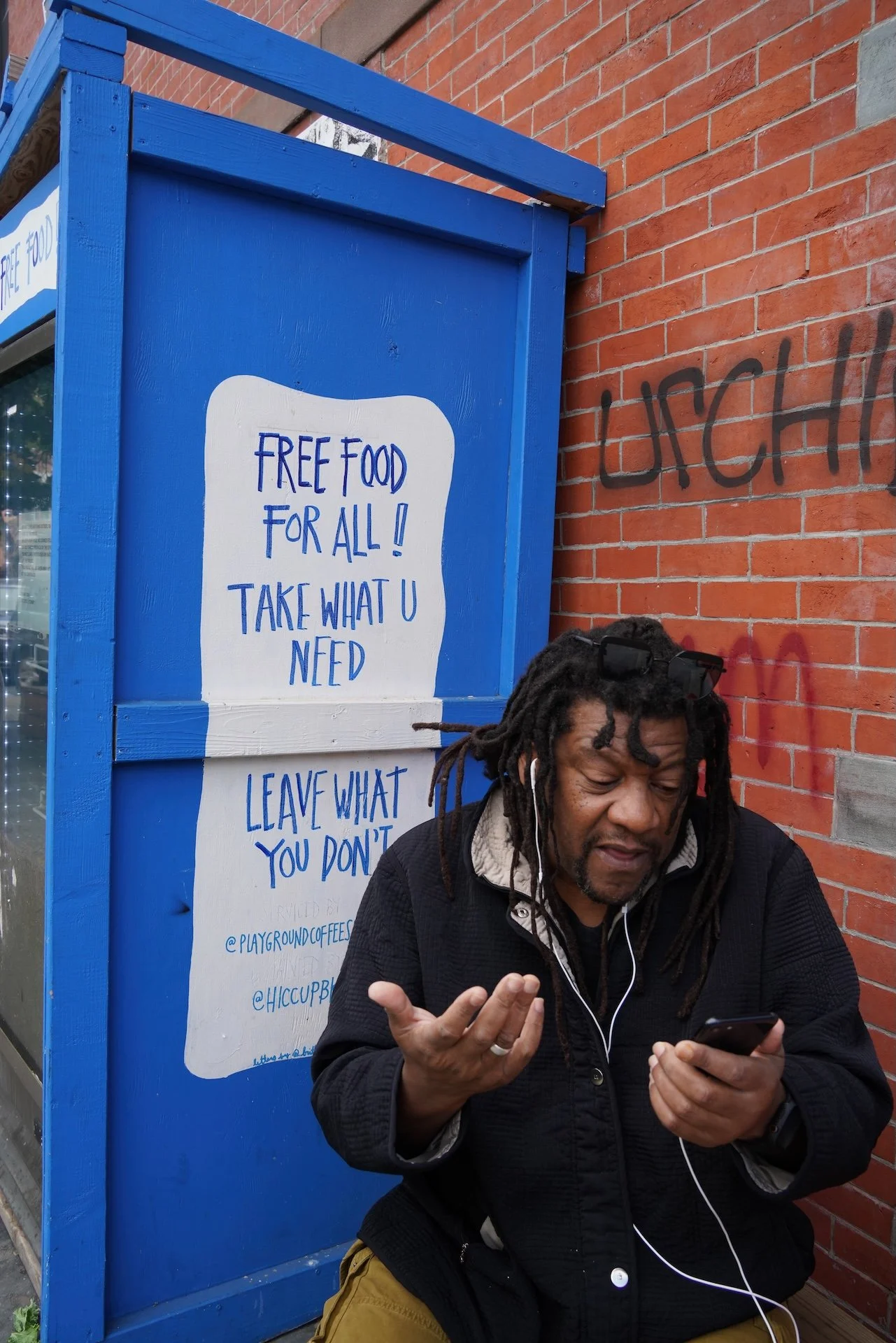 A man with dreadlocks and a black jacket standing in front of a blue wooden food sharing booth, holding a phone and making a hand gesture. The booth has a sign that reads 'FREE FOOD FOR ALL! TAKE WHAT U NEED' and is positioned next to a brick wall with graffiti.