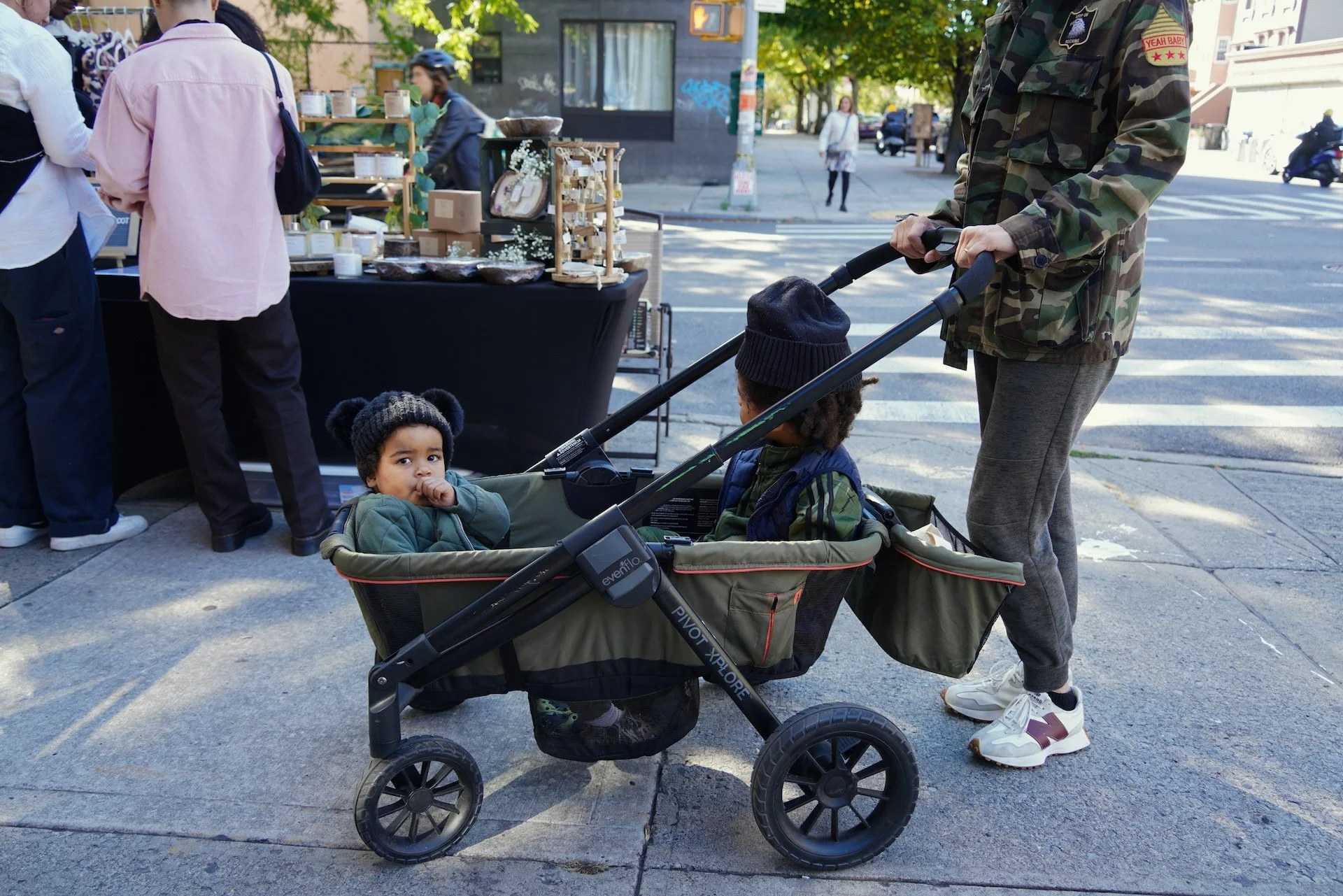 A young boy with curly hair and a green jacket sitting in a stroller, sucking his thumb and looking at the camera. An adult, partially visible, standing next to the stroller, wearing gray joggers, white sneakers, and a camouflage jacket. The scene is on a city sidewalk with a street and a crosswalk visible in the background. A woman in a white shirt and another woman in a pink shirt are near a street vendor stand displaying various jars and boxes.