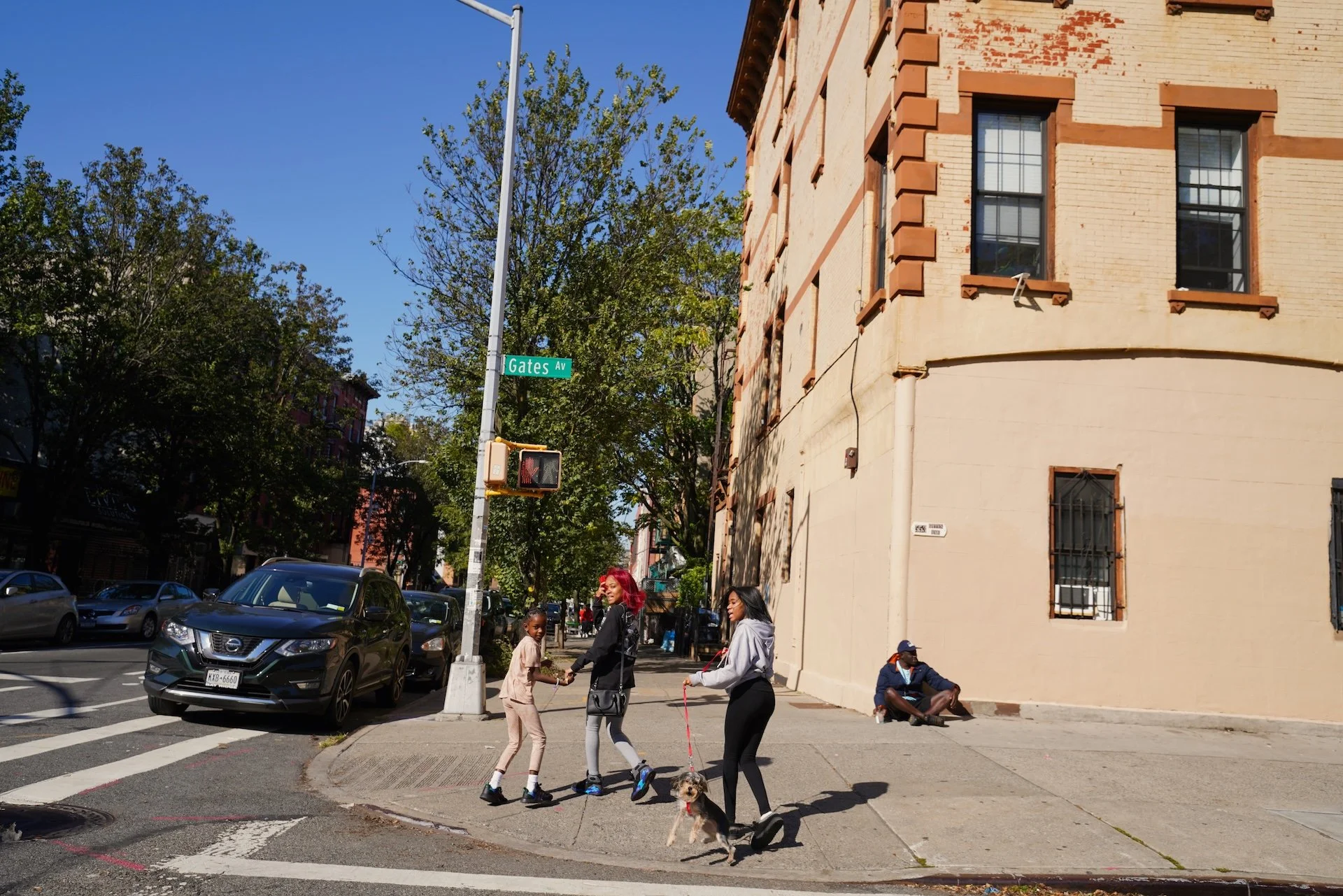 People crossing the street at the corner of a city block on Gates Avenue. The street corner has a traffic light, street sign, and parked cars. There is a large beige brick building with windows, and trees with green leaves in the background. Several people, including a woman with bright red hair, are walking, and some are sitting on the sidewalk.