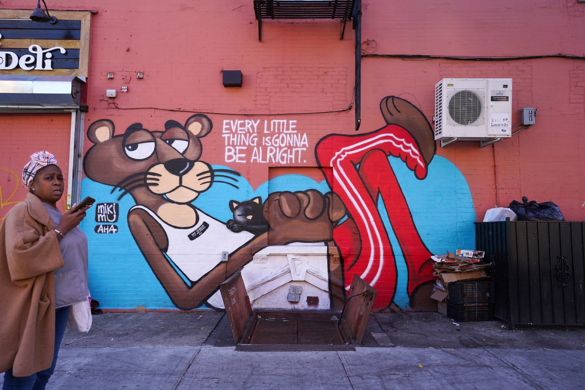 Street mural of a cartoon bear with a black cat, accompanied by the text "Every little thing is gonna be alright." A woman in a tan coat and headscarf is standing nearby, looking at the camera. The mural is on a red brick wall with an air conditioning unit and some garbage bags nearby.