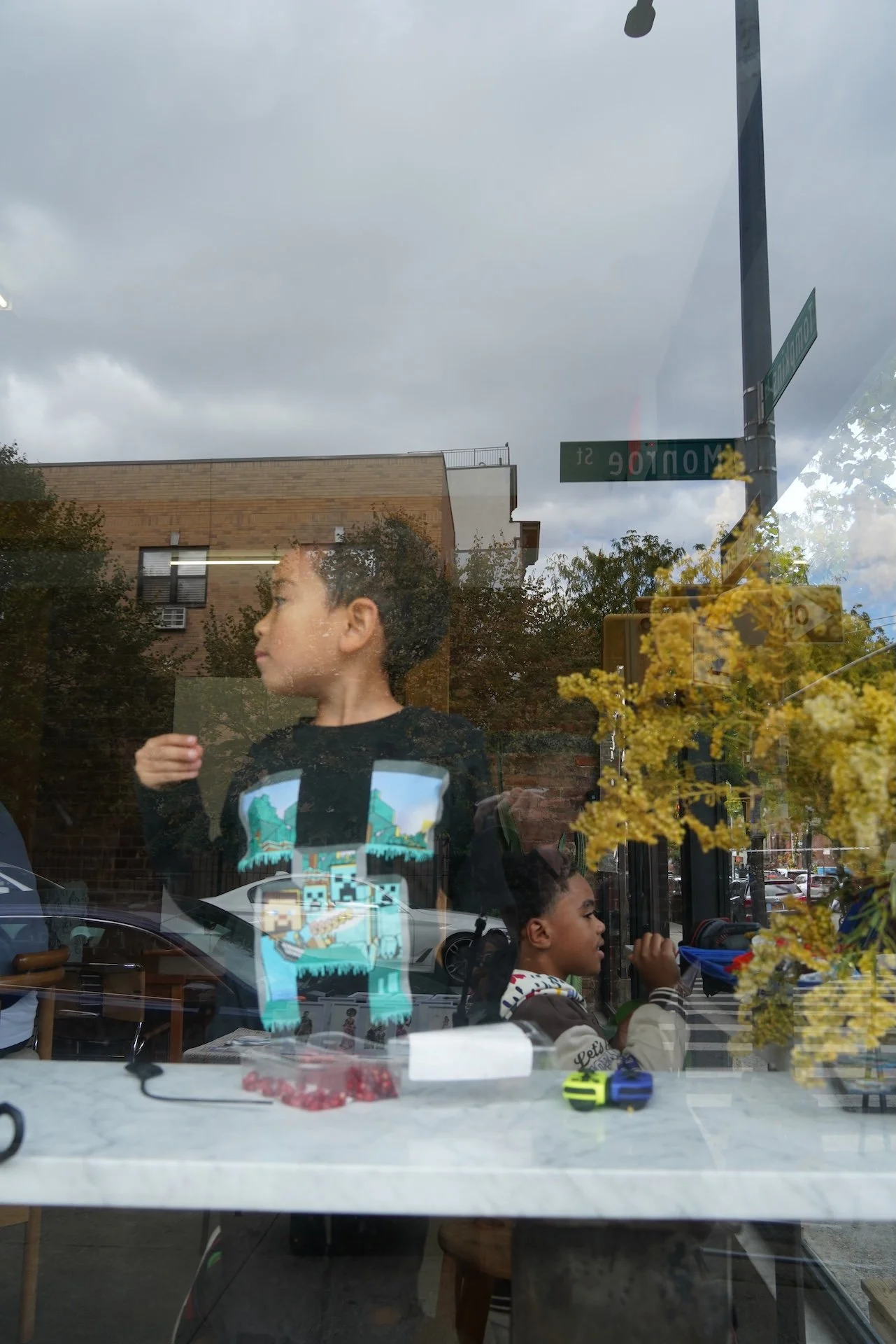 The photo is taken through a window showing a city street with pedestrians, trees, and a brick building under a cloudy sky. Inside, there are children sitting at a table, one with a video game on his shirt, and a girl with braids talking to someone off-camera. The reflection of street signs and yellow flowers is visible on the glass.