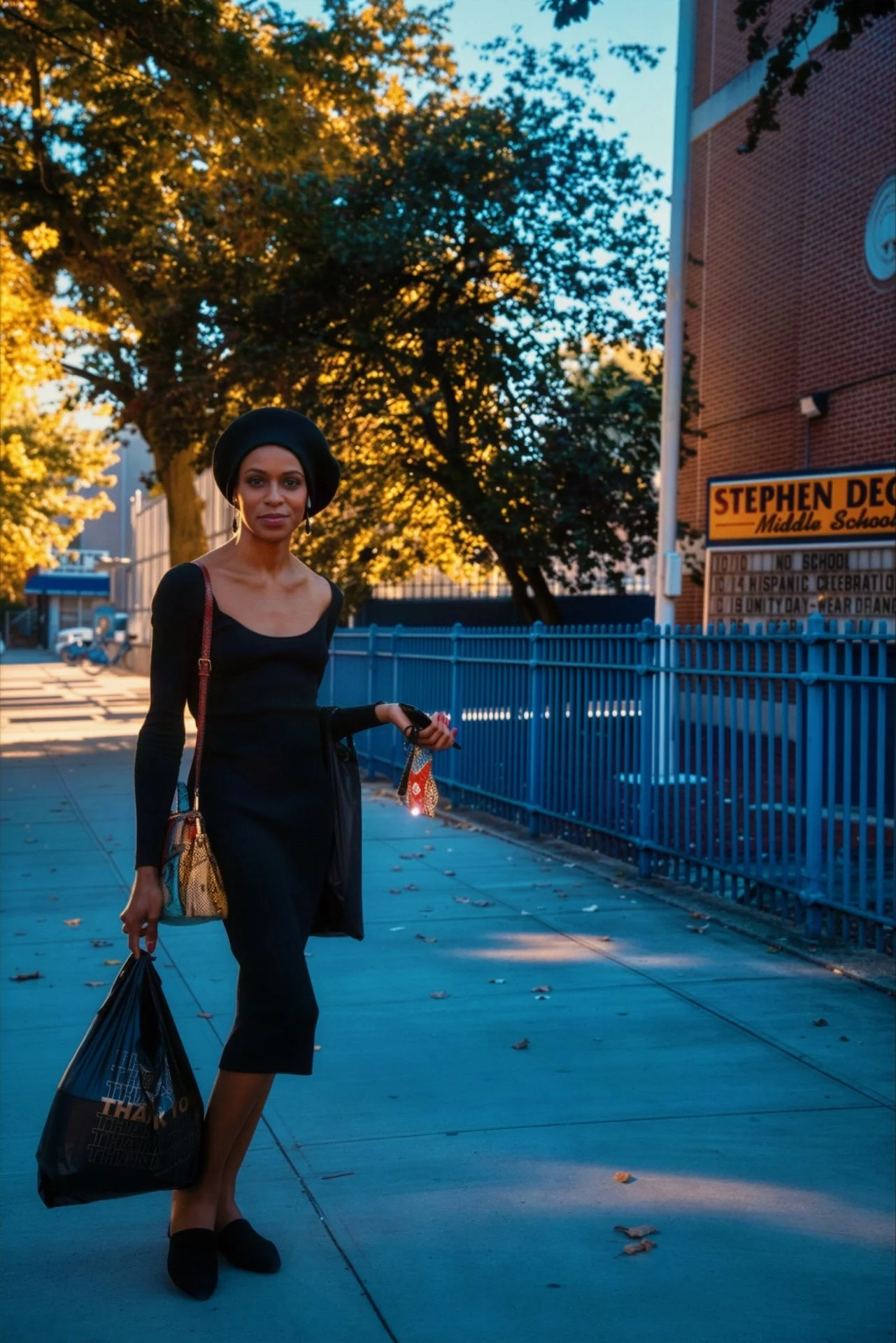 A woman dressed in black, wearing a hat, standing on a sidewalk in front of a blue fence and a brick school building. She is holding a shopping bag and a small item, with trees and a clear sky in the background during sunset.