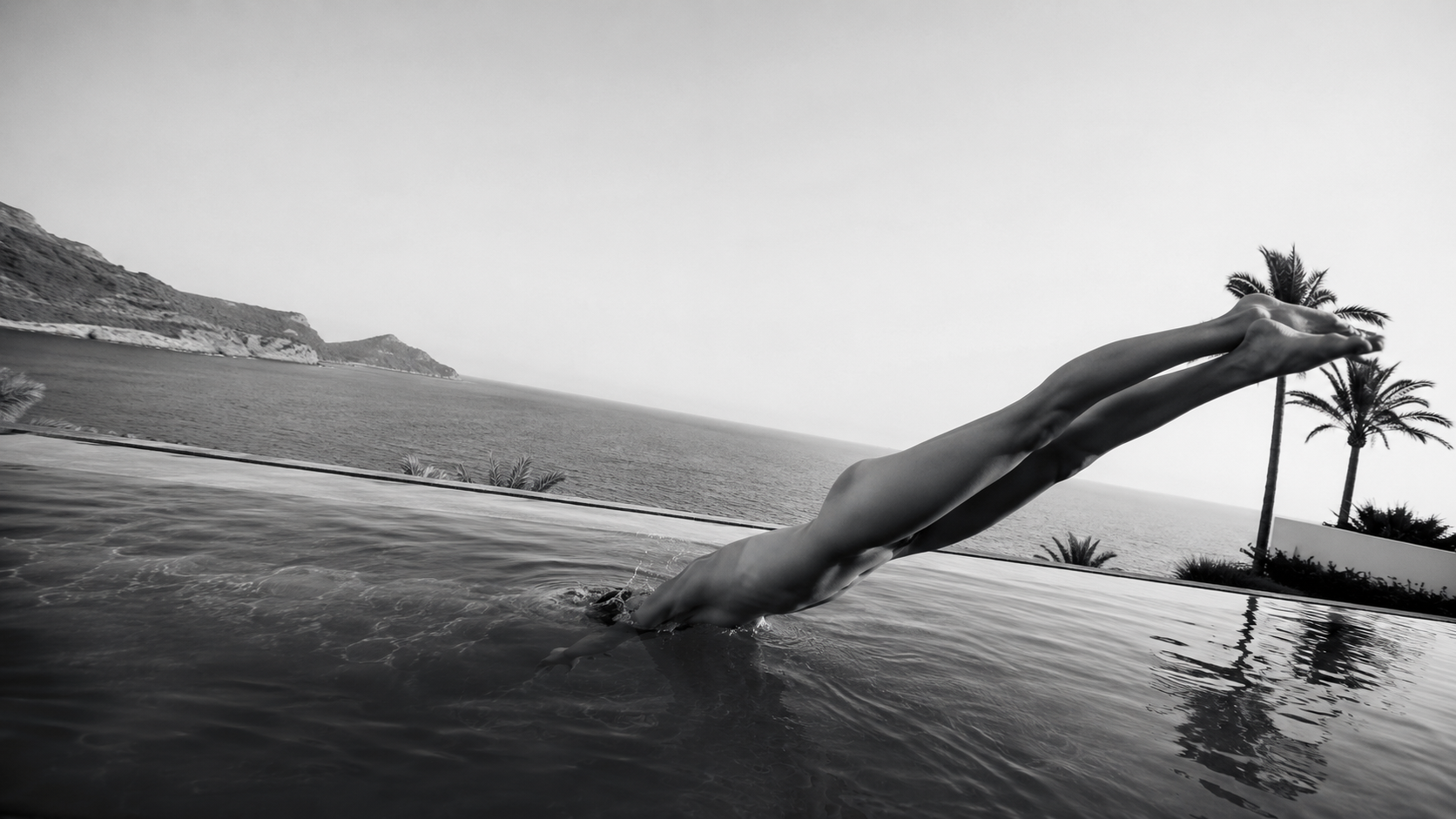 Person diving into a pool with a scenic ocean view, palm trees, and hills in the background in black and white.