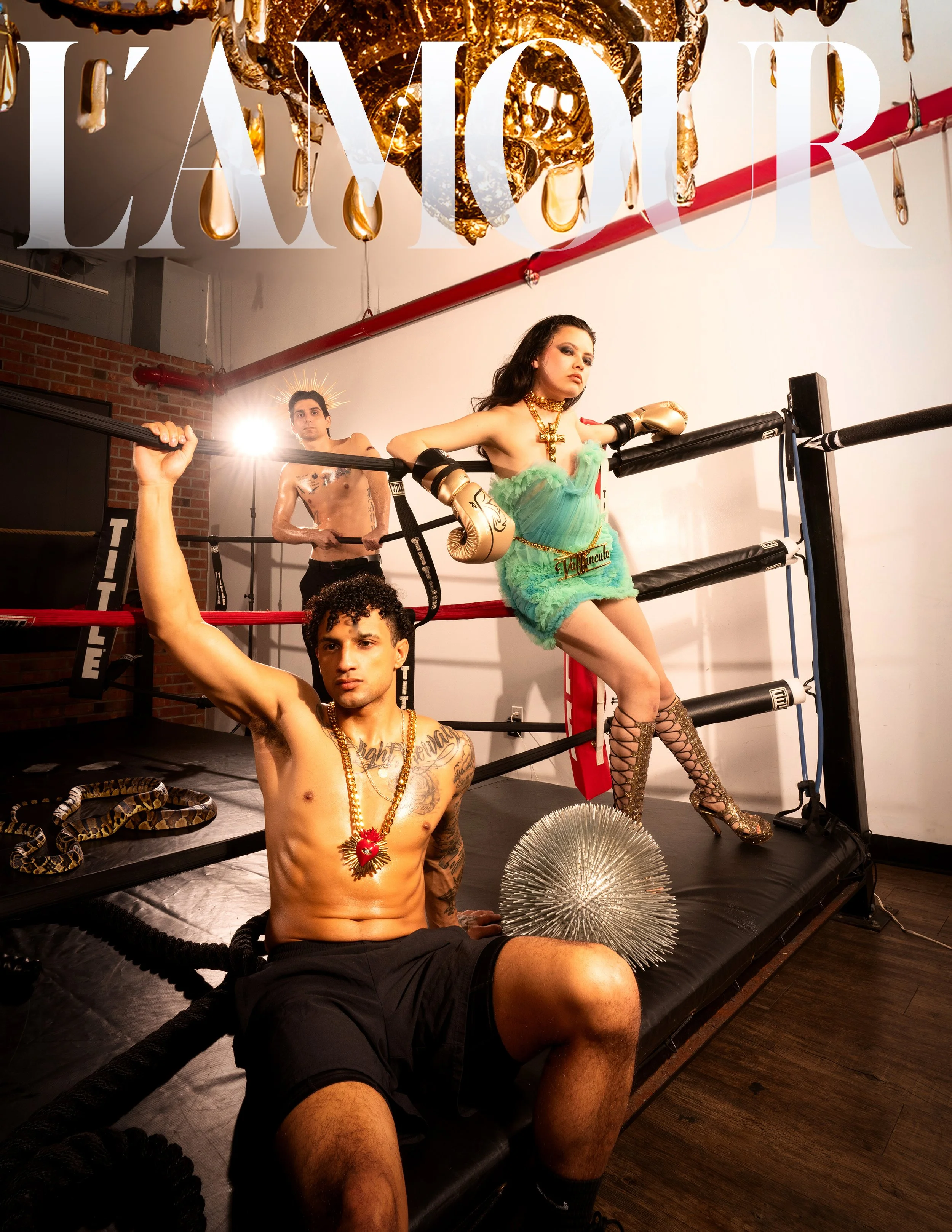 Three models in a boxing ring with a chandelier, snakes, and fashion attire, under the text 'L'AMOUR'.