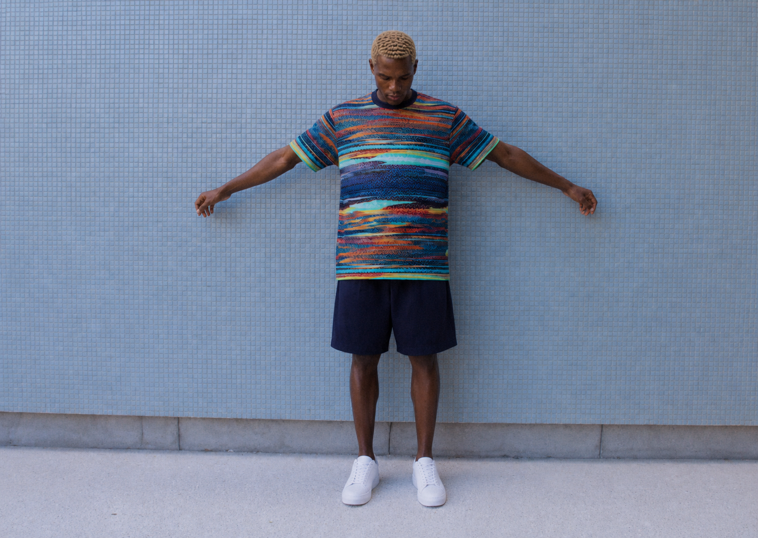 THE OUTNET AI fashion campaign in São Paulo created by Alessia Moccia, featuring a man in a colorful knit shirt and navy shorts against a blue tiled wall.