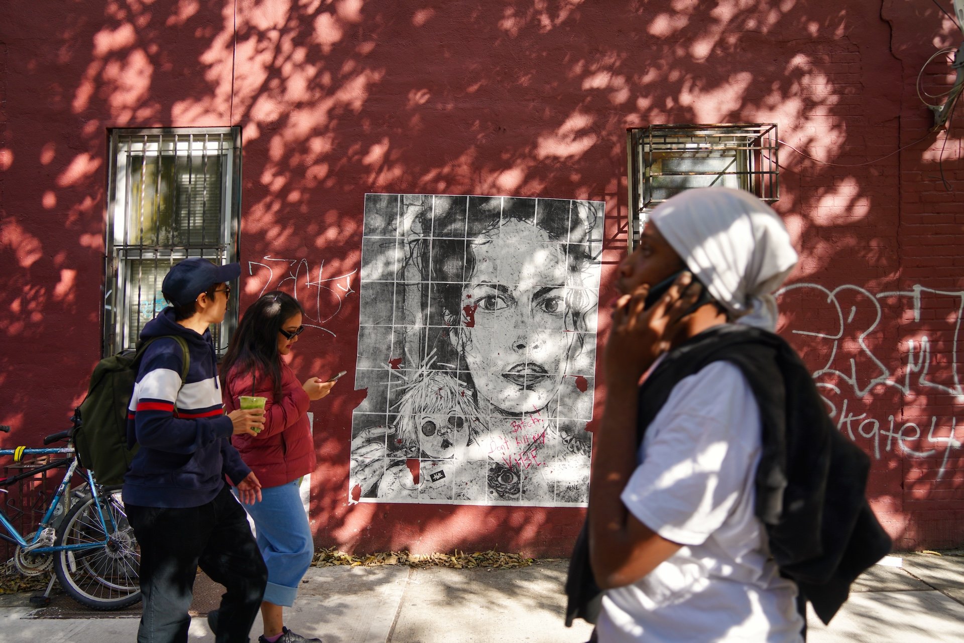 Street scene with three people walking past street art on a red brick wall, shadow of tree branches overhead, two windows with metal bars, and a bicycle leaning against the wall.