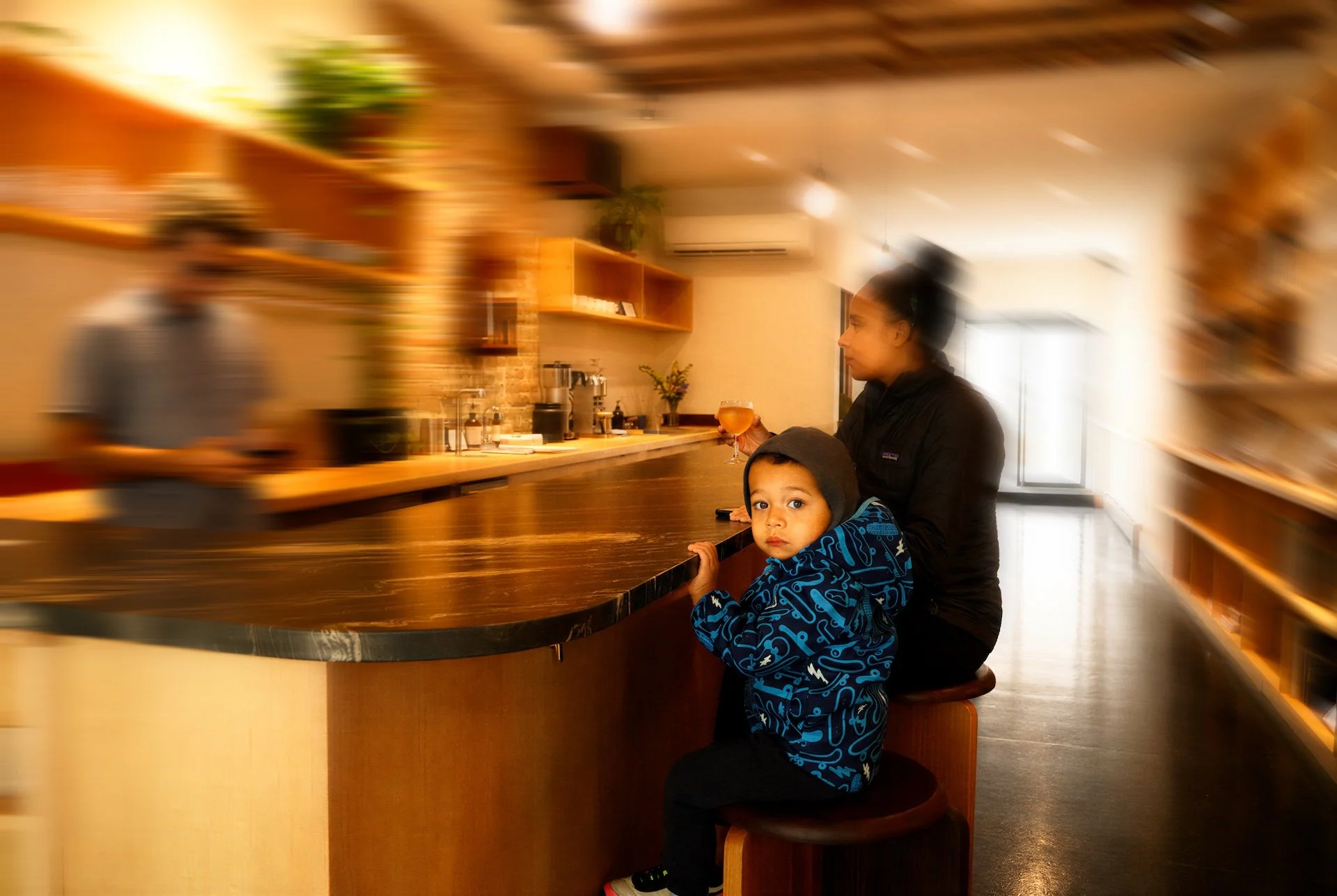 A woman and a young boy sit at a bar counter inside a cozy cafe with warm lighting and wooden shelves. The woman is holding a drink and appears to be waiting, while the boy looks directly at the camera with a curious expression.