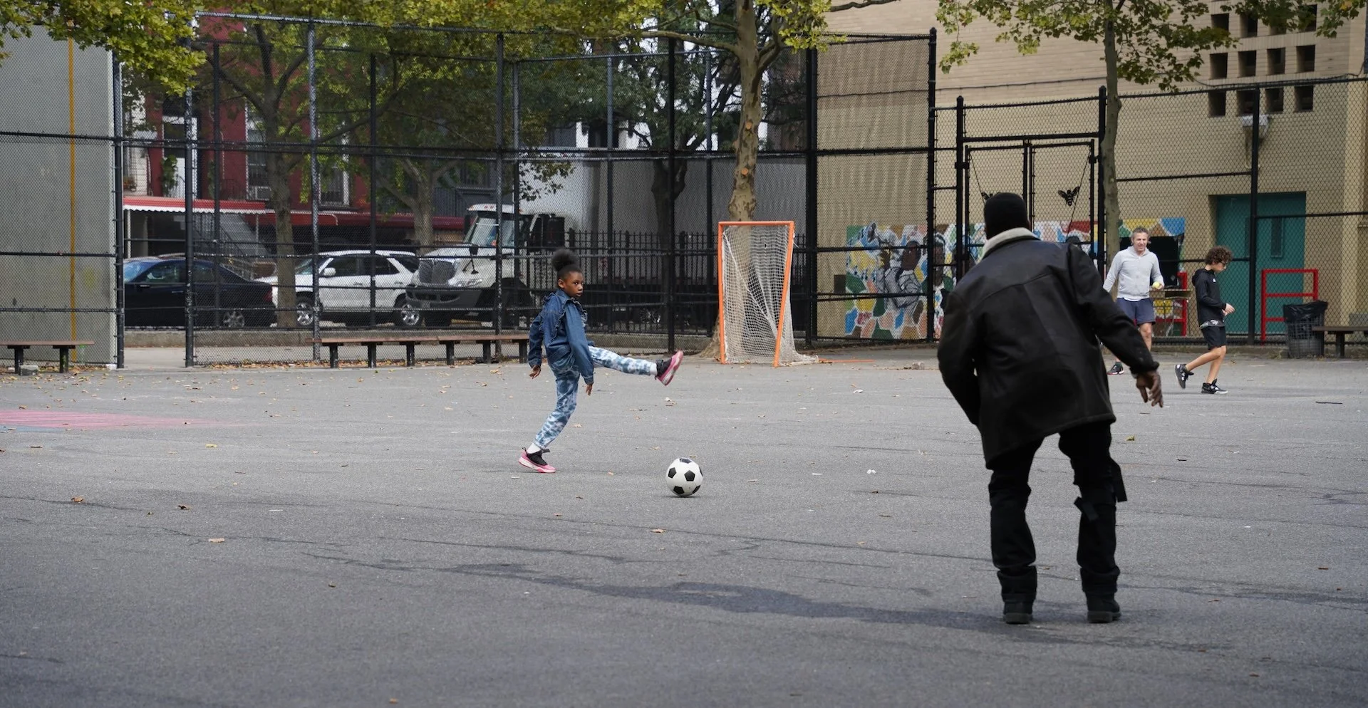 Children playing soccer on an outdoor court with a chain-link fence, surrounded by parked cars and buildings, during daytime.