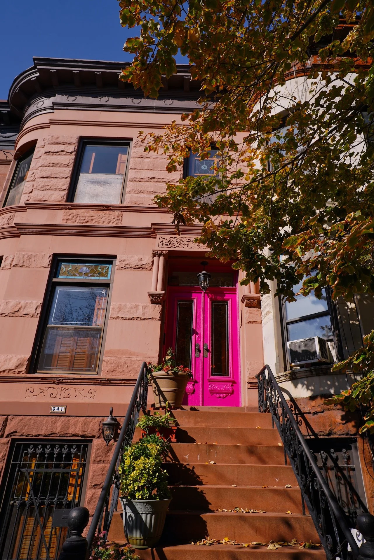 A brownstone building with a vibrant pink front door, black iron stairs, and balcony, surrounded by leafy trees under a clear blue sky.