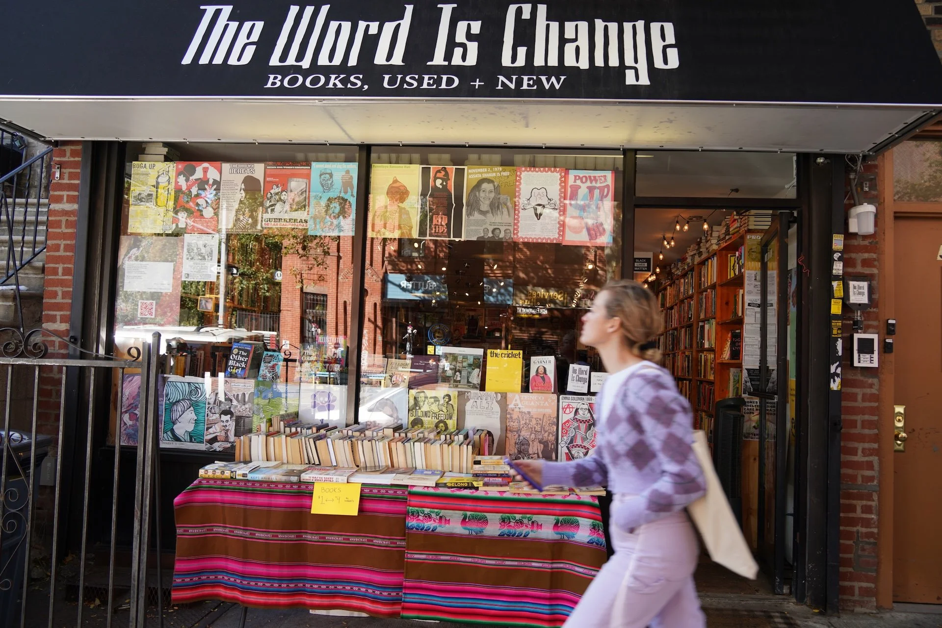 A bookstore storefront named "The Word Is Change" with a sign indicating they sell used and new books. The display window showcases various colorful posters and books. In front, a table with a vibrant, striped tablecloth holds a selection of books for sale, with a yellow sign indicating prices. A woman in a light purple tie-dye hoodie is walking past the store, holding a book or notebook, with the interior of the shop visible behind her.