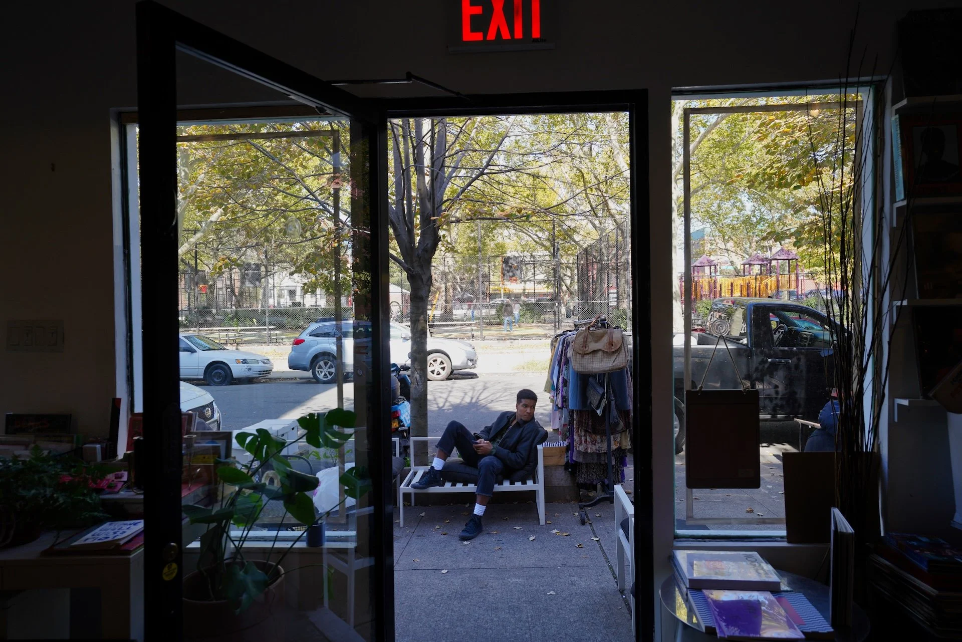 View from inside a store looking out onto a sidewalk with cars parked on the street. There are two people sitting outside on a bench, with clothing hanging nearby and trees in the background. An illuminated red 'EXIT' sign is visible at the top of the door frame.