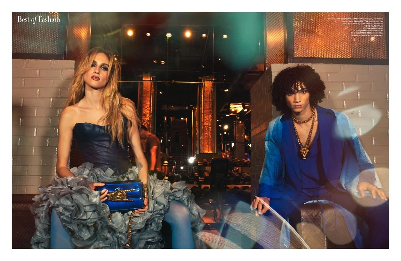Fashion photoshoot featuring two models in stylish outfits, one in a strapless dress with a ruffled skirt and holding a blue handbag, and the other in a blue outfit with a gold necklace, set in a dimly lit interior with decorative lighting.