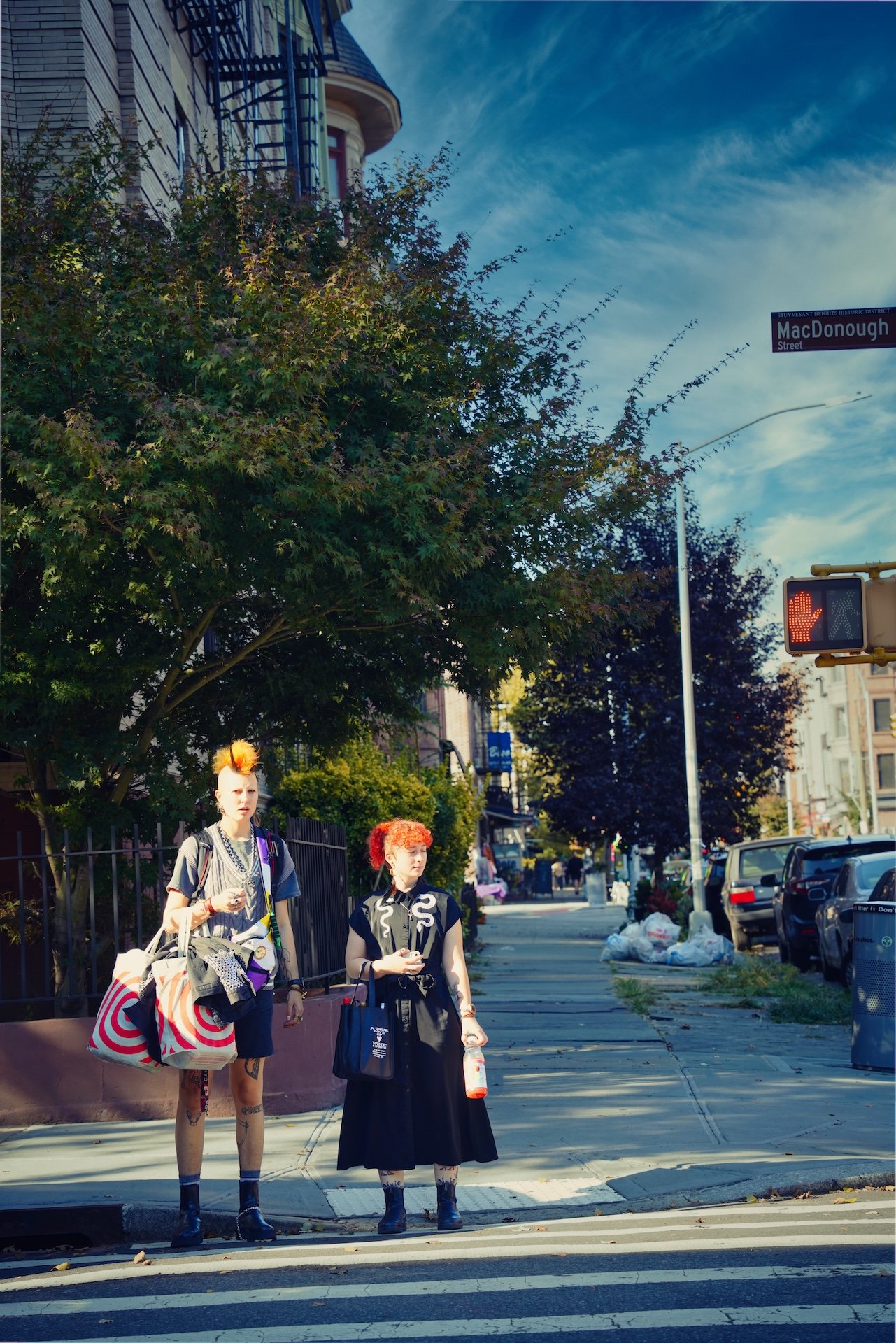 Two women waiting at a crosswalk on a city street with trees, cars, and a building in the background. One woman has a mohawk hairstyle with orange and yellow hair, wearing a gray outfit with tattoos on her legs, carrying shopping bags. The other woman has curly red hair, wearing a black dress with white designs, holding a water bottle. Traffic light shows a red hand signal indicating pedestrians should not cross.