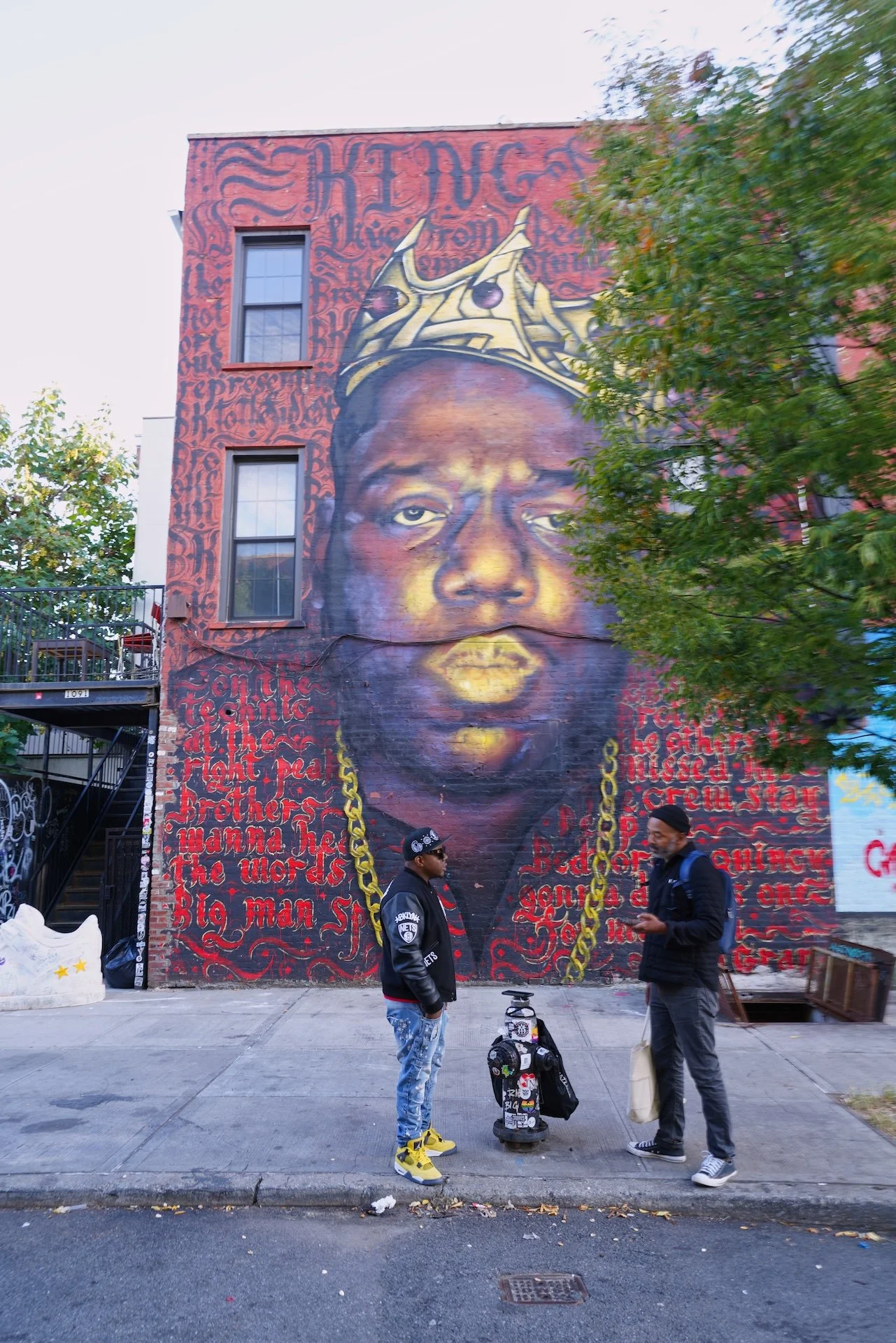 Street scene with a large mural of a person's face on a brick building, featuring detailed graffiti and red text backgrounds. Two people are standing in front of the mural, one in a Brooklyn Nets jacket and the other reading a phone. The sidewalk has some debris and trees partially cover the mural.