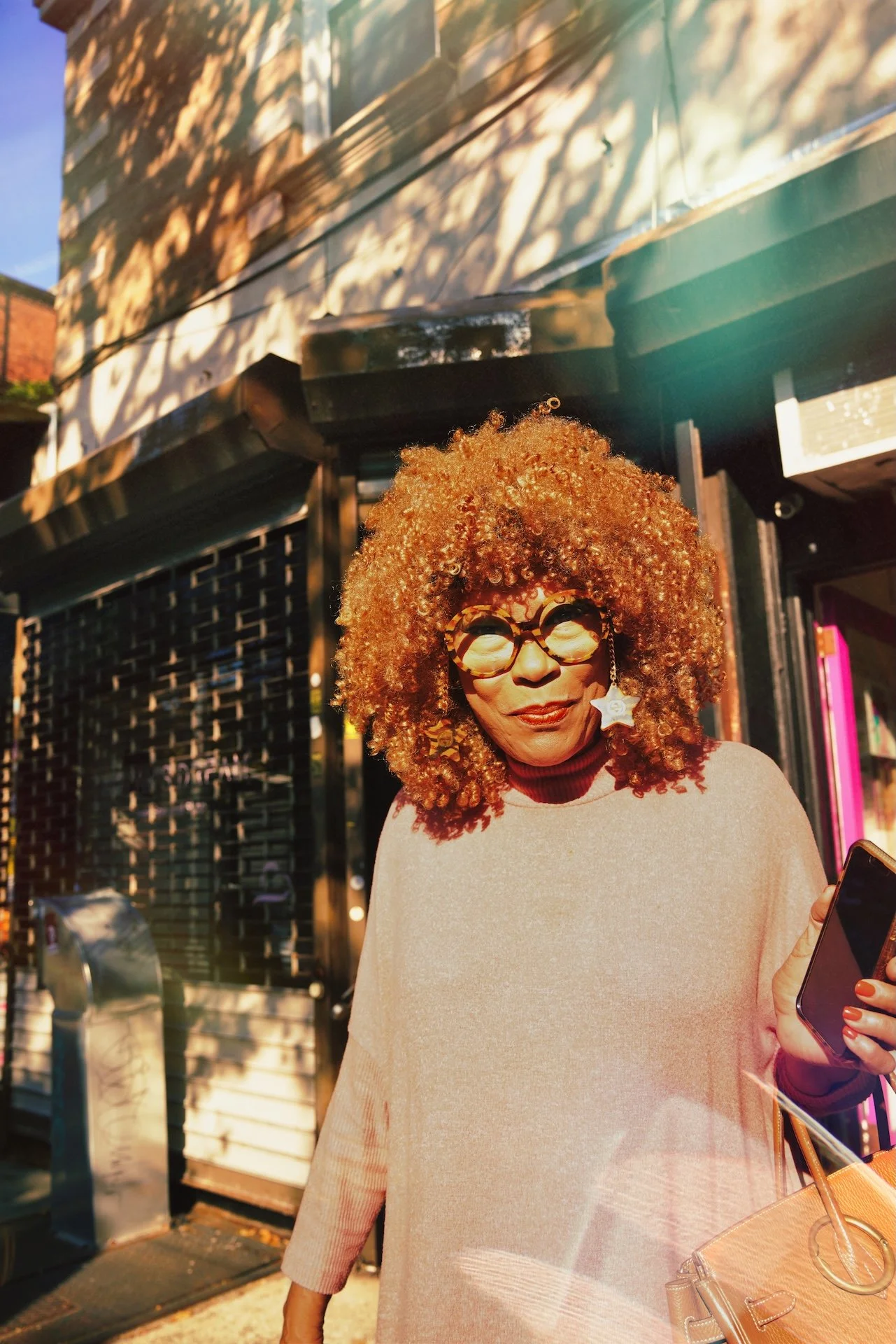 A woman with curly red hair and tortoise shell glasses standing outside in the sunlight, wearing a beige sweater and carrying a beige bag, with shadows from trees on the building behind her.