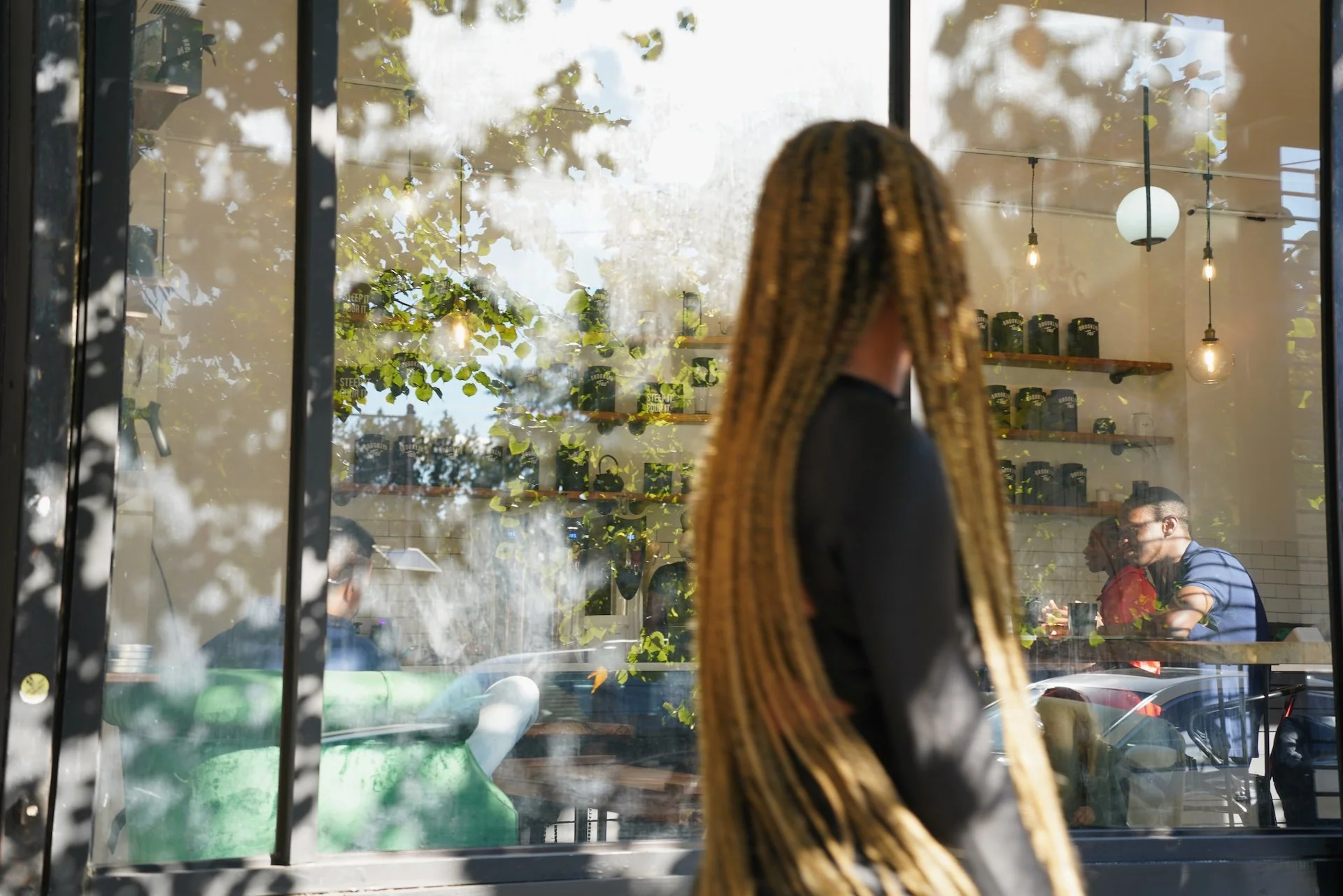 People sitting inside a cafe, viewed through a window with reflections of trees and outdoor scenery, warm lighting, and shelves with jars displaying. Someone with braided hair is standing outside.
