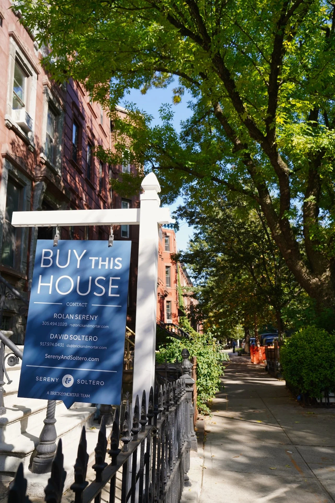 A sidewalk view on a sunny day in an urban neighborhood with trees, brownstone buildings, and a sign that says 'Buy This House' from a real estate team.