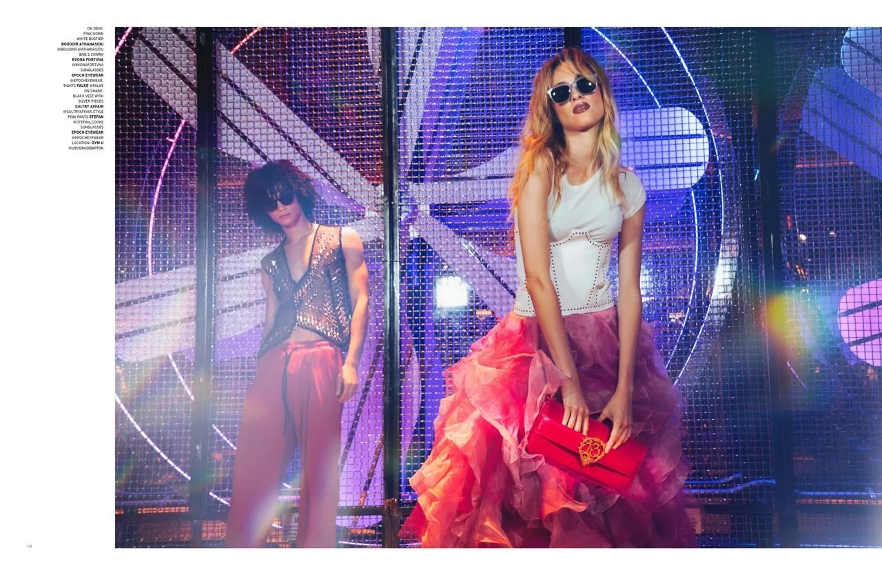 Fashion models in vibrant nightclub setting with neon lights, one wearing a chain top and red pants, the other in a white bodice with a pink skirt, holding a red clutch.