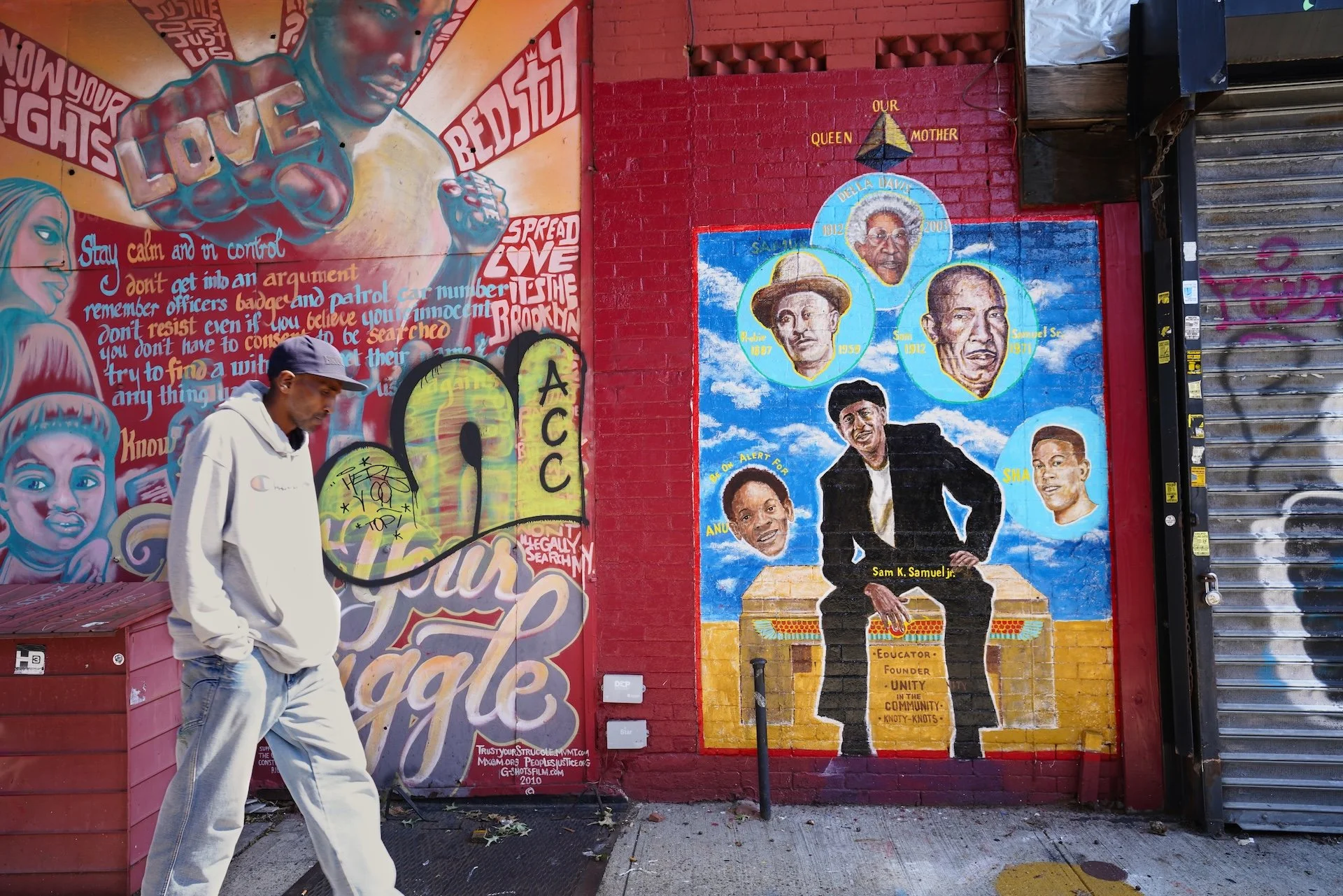 Street mural of influential African American figures with a man walking in front, wearing a gray cap and hoodie, on an urban sidewalk.