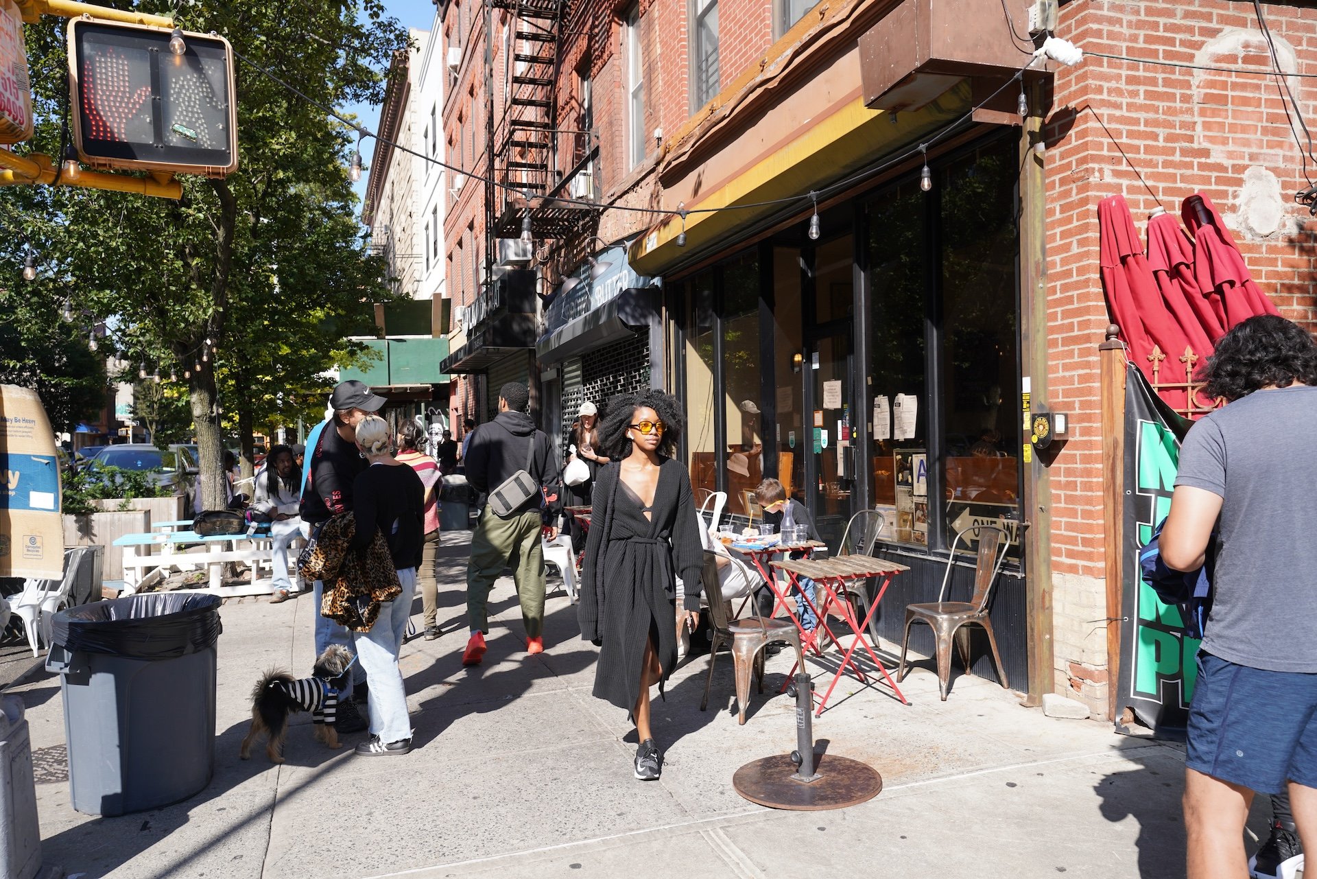 A busy city sidewalk outside a restaurant with people walking and sitting at outdoor tables. There are red umbrellas and a brick building with a fire escape. Some people are wearing casual and fashionable clothing, and a woman in a black dress and sunglasses is in the foreground.