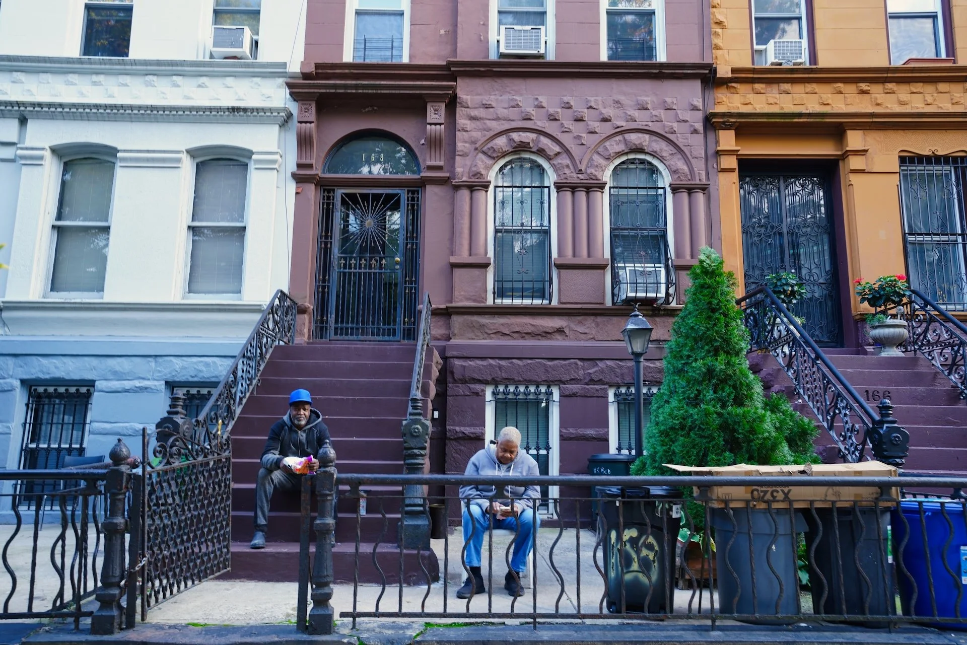 Three men sitting on the steps in front of brownstone houses on a city street. The man on the left is wearing a blue hat and black jacket, holding a snack. The man in the middle is wearing a gray hoodie, looking down, possibly at a phone. The man on the right is wearing a gray hoodie, looking down at something in his hands. A black wrought-iron fence surrounds the area, and there is a small tree and a street lamp on the sidewalk.