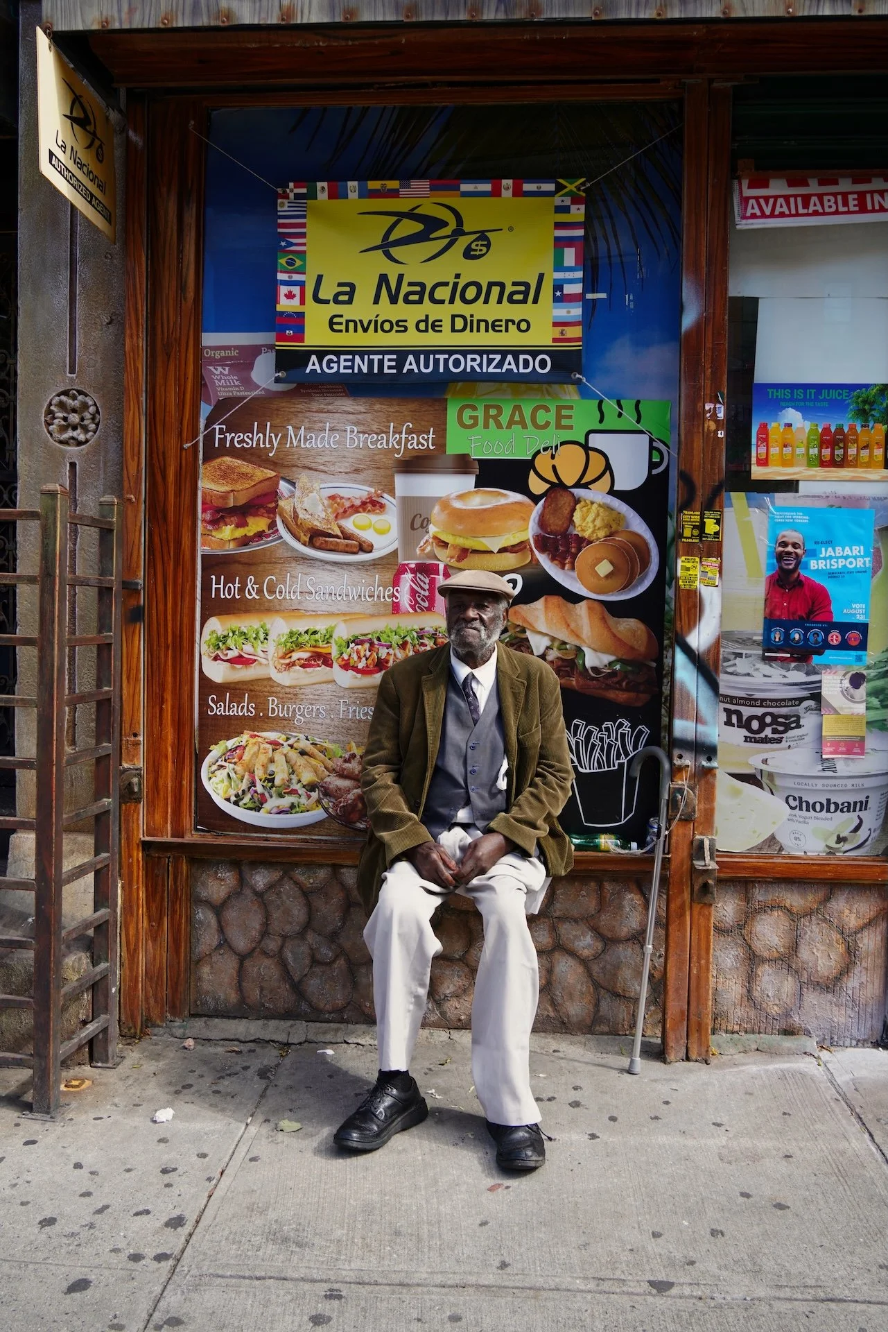 An elderly man with gray hair and beard sitting on a sidewalk in front of a restaurant, wearing a brown jacket, light-colored pants, and black shoes, holding a walking cane, with a large colorful food menu behind him.