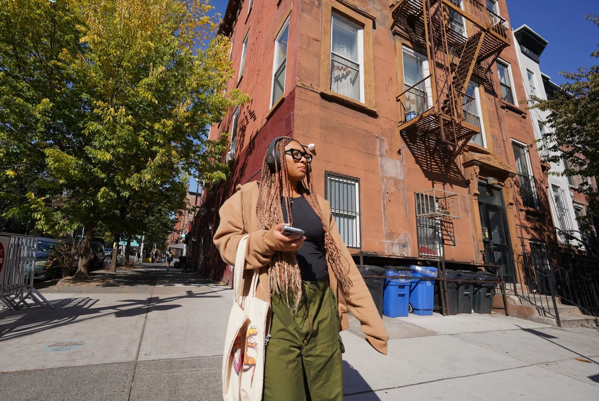 A woman with long braided hair and glasses walking on a city sidewalk, holding a smartphone, with a brown brick building with fire escape stairs, green trees, parked cars, and blue recycling bins in the background on a sunny day.