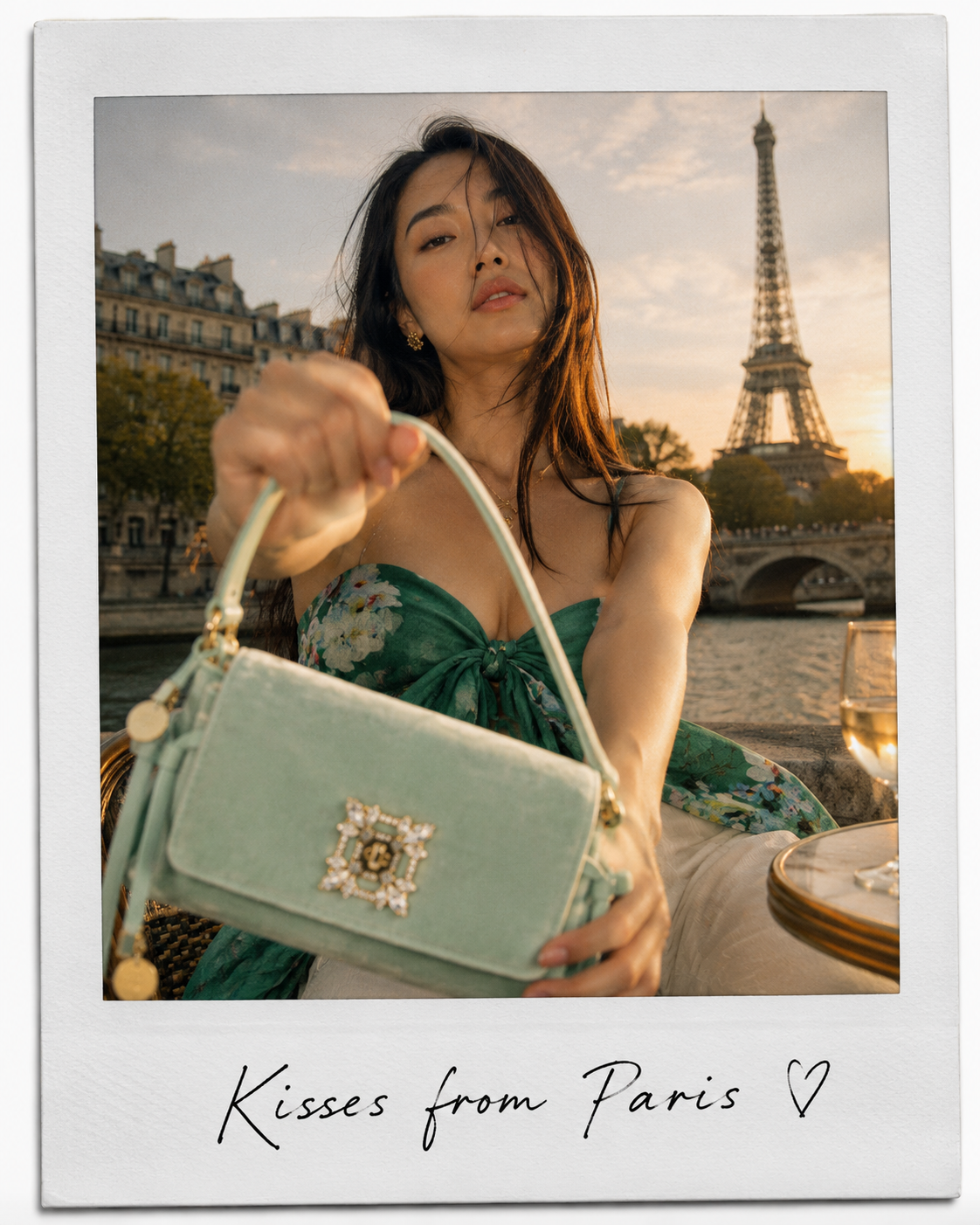 A woman holding a light green handbag with a decorative jewel clasp, sitting outdoors near the Seine River with the Eiffel Tower in the background during sunset, with a glass of white wine nearby. The photo has a handwritten note at the bottom that says "Kisses from Paris" with a heart.