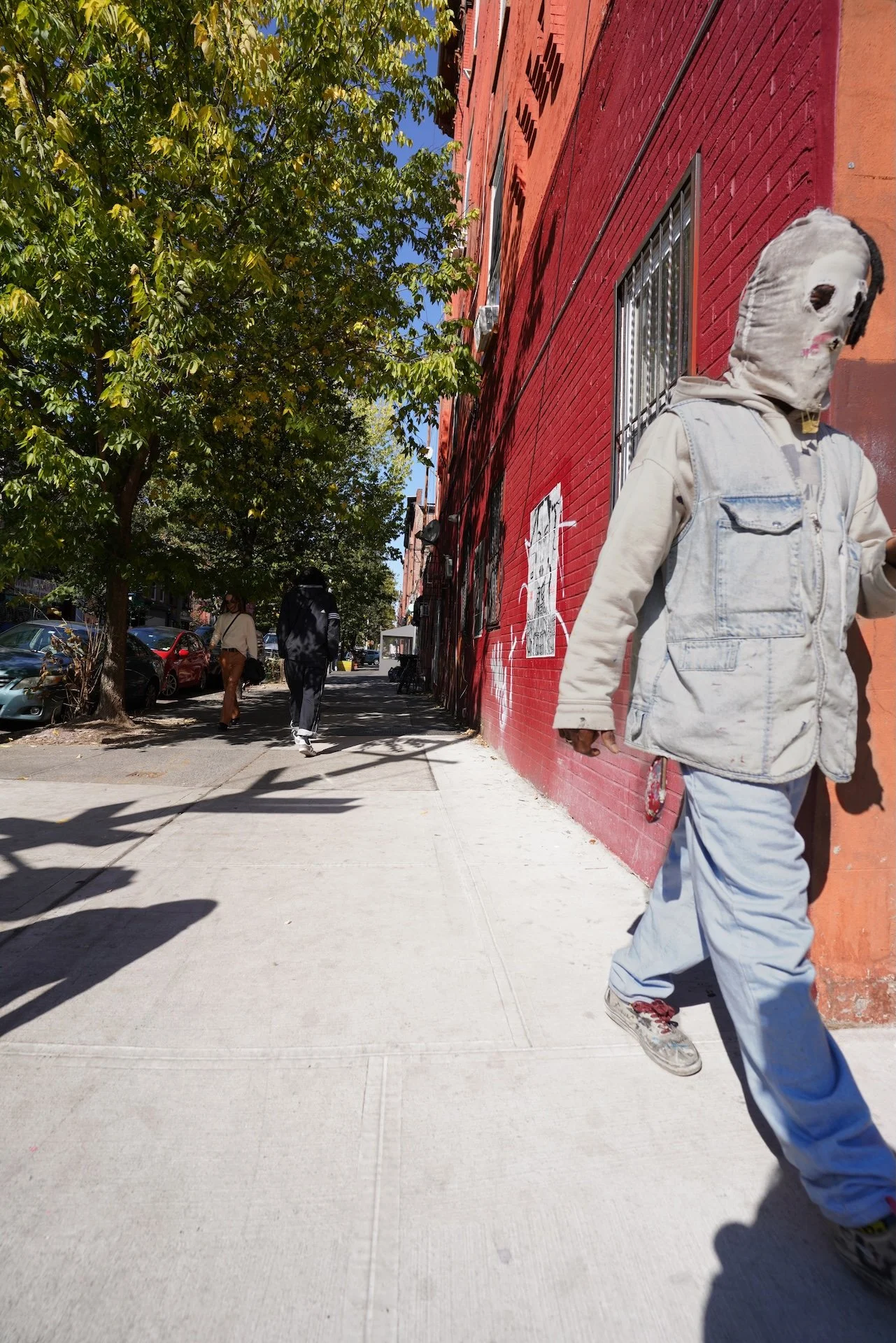 People walking along a sidewalk next to a red brick building with graffiti, and a tree with green leaves shading part of the sidewalk on a sunny day.