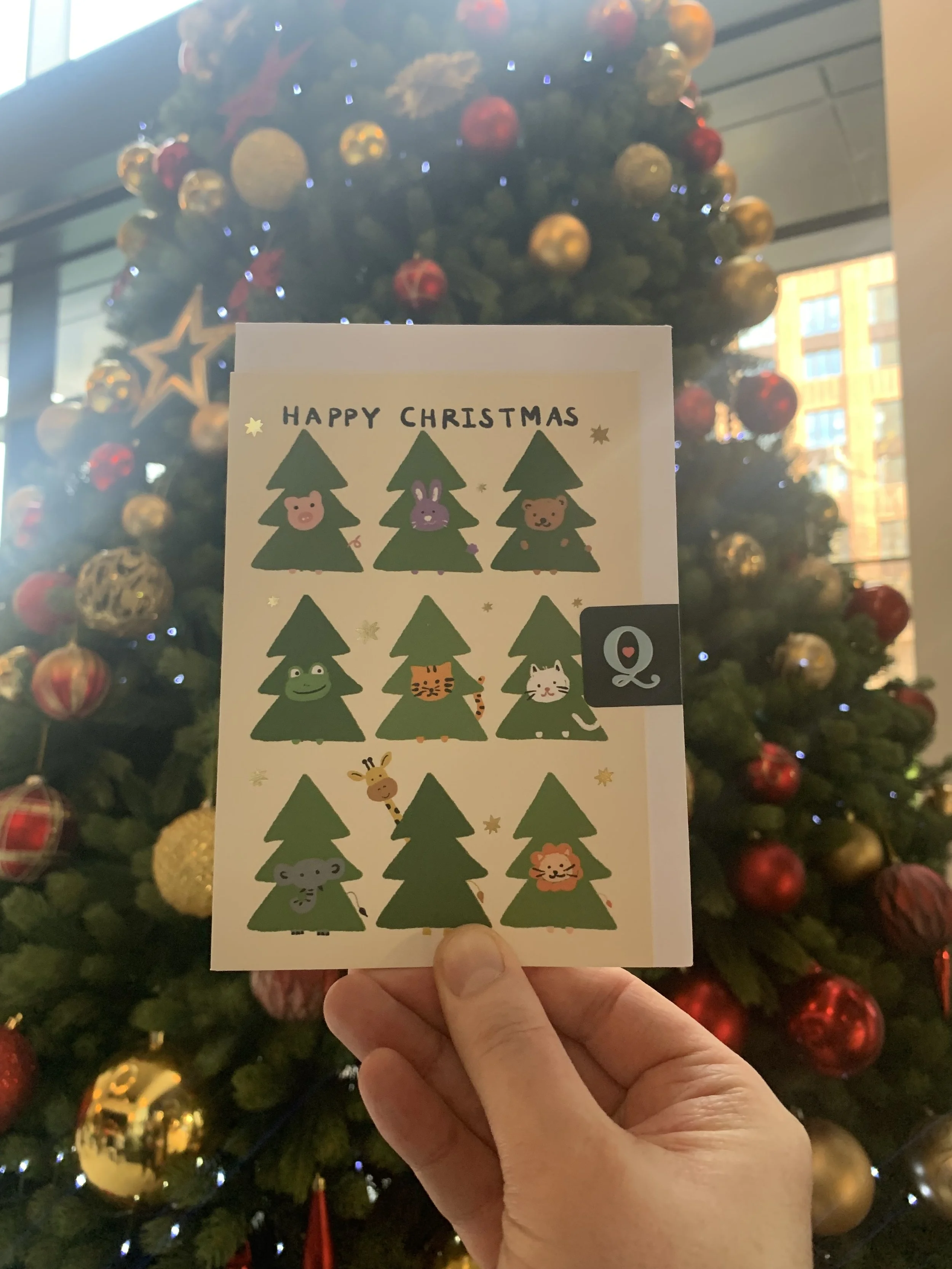 A hand holding a Christmas card featuring cartoon animals each poking their heads out of Christmas trees, and held up against a decorated Christmas tree in the background.