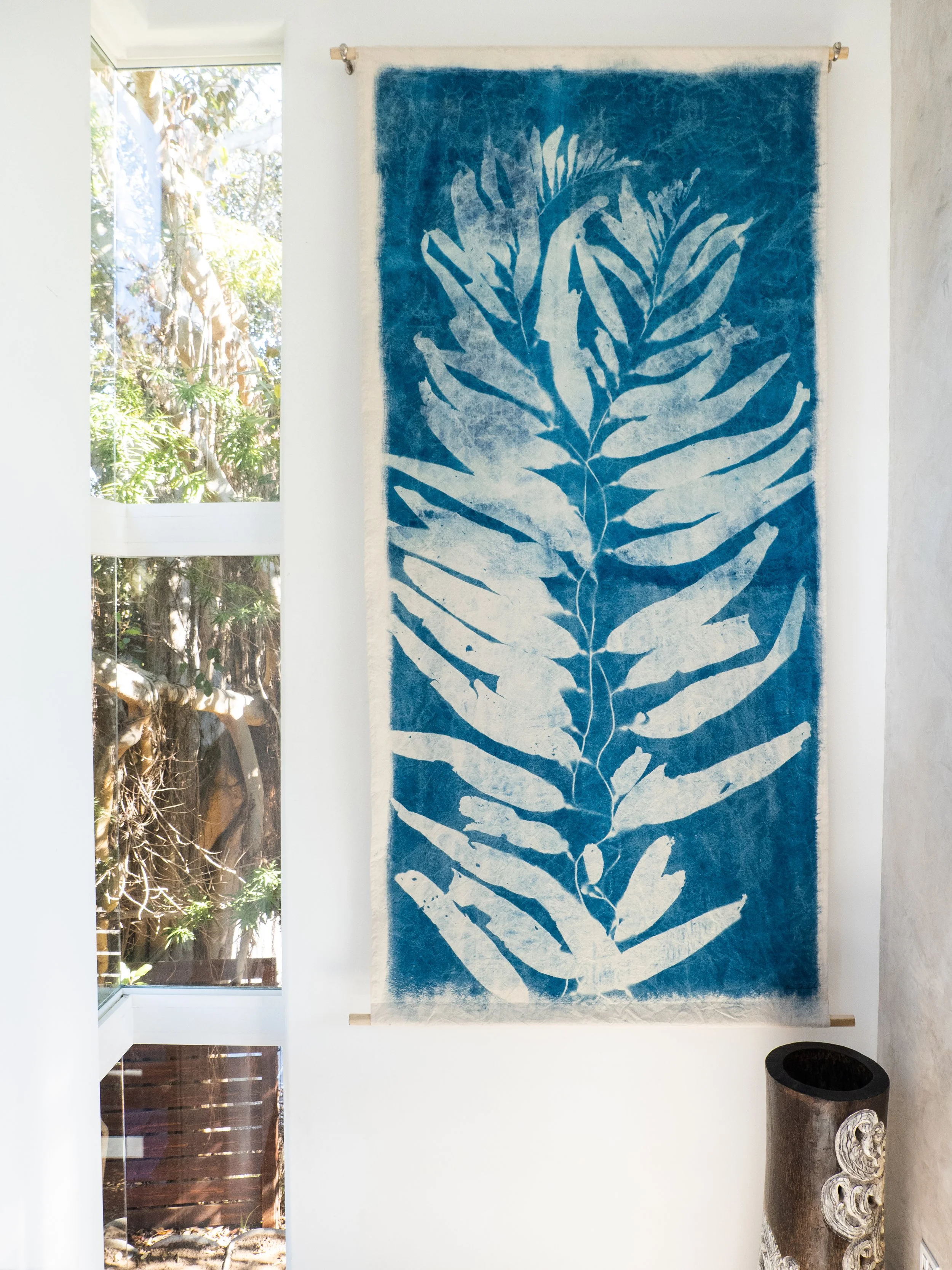 A tall, vertical textile wall hanging with a blue background and a white leaf pattern, mounted on a wall beside a window showing trees outside.