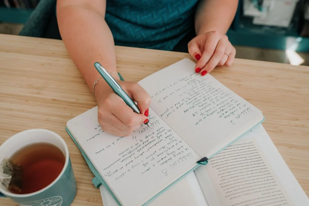 A person with red painted nails is writing in a notebook with a teal pen. There is a paper with printed text underneath the notebook and a cup of tea with a tea bag on a wooden table.